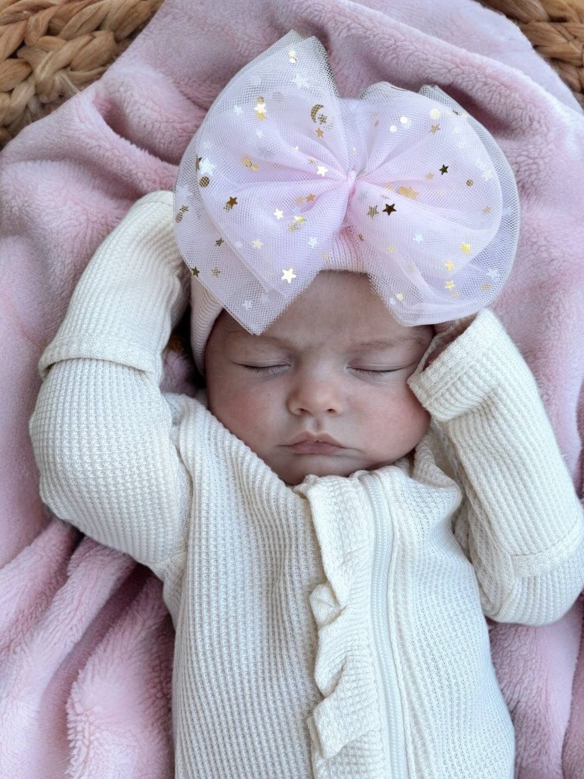 Baby girl sleeping with a large pink bow and white outfit, lying on a pink blanket.