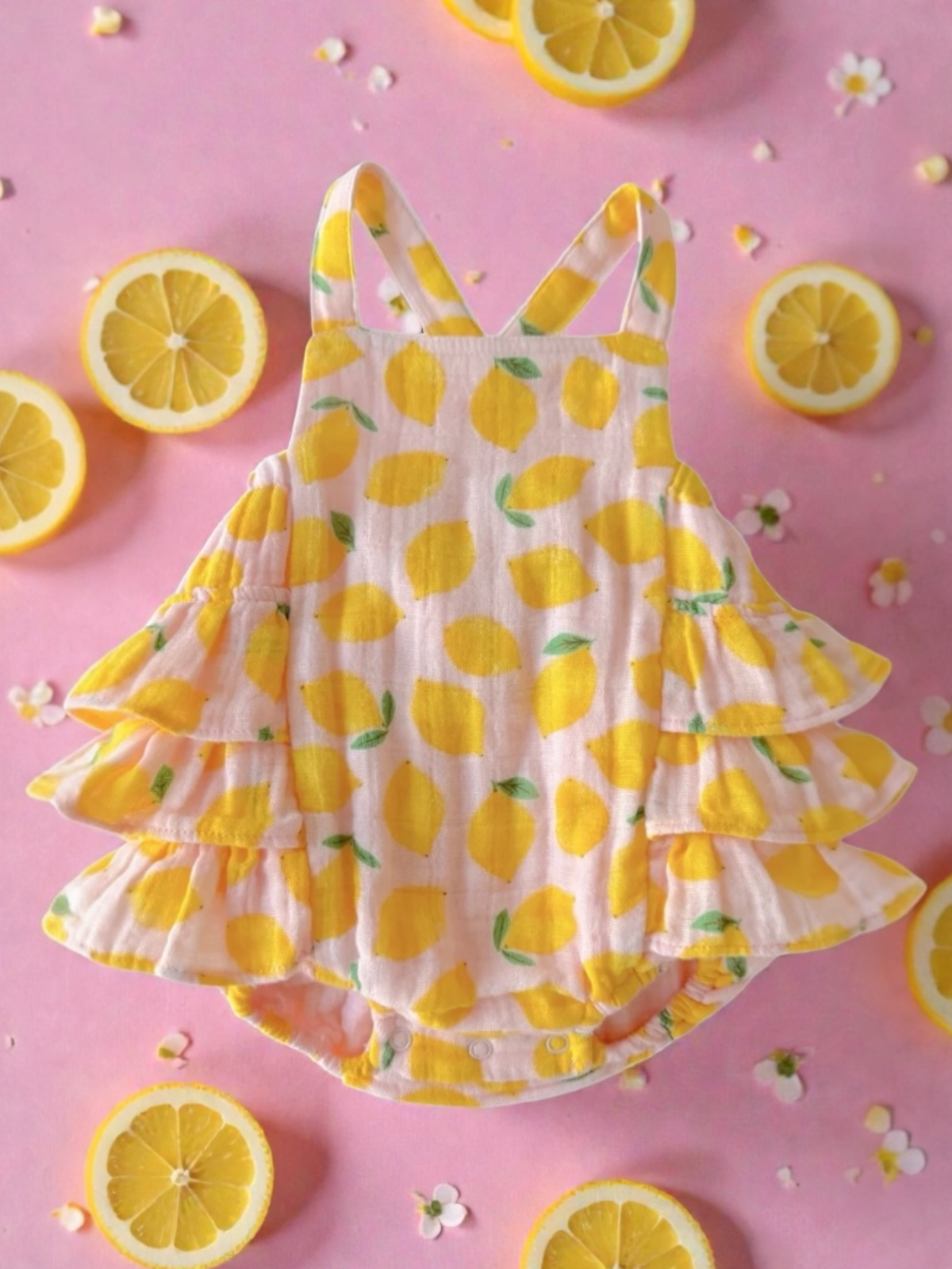 Yellow lemon-patterned baby romper with ruffle sleeves on a pink backdrop adorned with lemon slices and flowers.