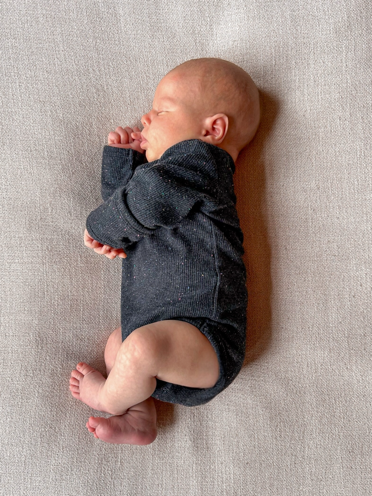 Newborn baby peacefully sleeping on a soft surface, wearing a dark onesie, with hands near the face.