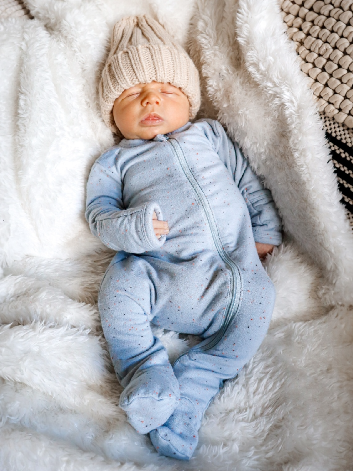 Newborn baby sleeping in a cozy blue onesie and cream knit hat on a fluffy blanket.
