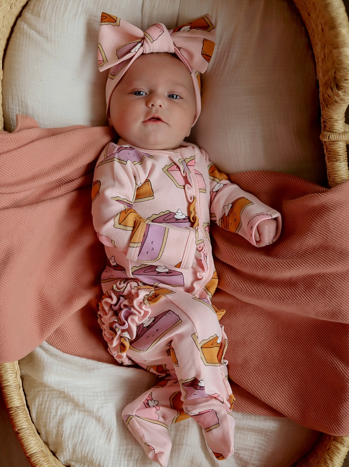 Baby in a pink outfit with dessert patterns, wearing a matching headband, lying on a soft, textured blanket.