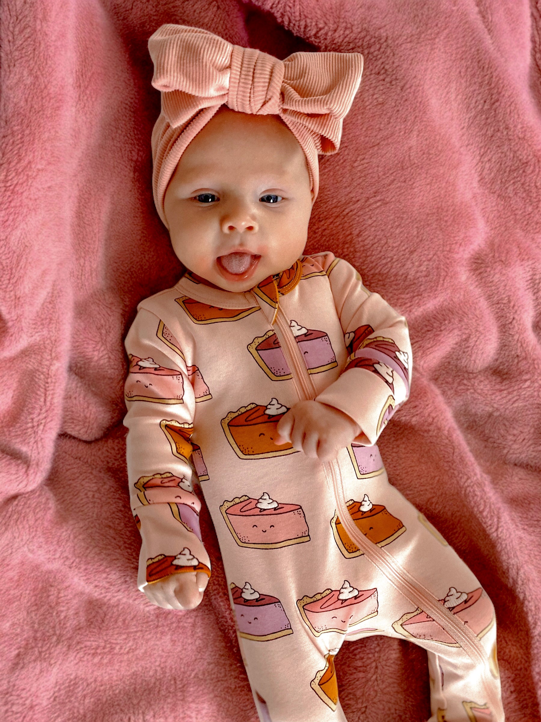 Baby in pink dessert-patterned outfit and large bow, lying on a soft pink blanket, sticking out tongue.