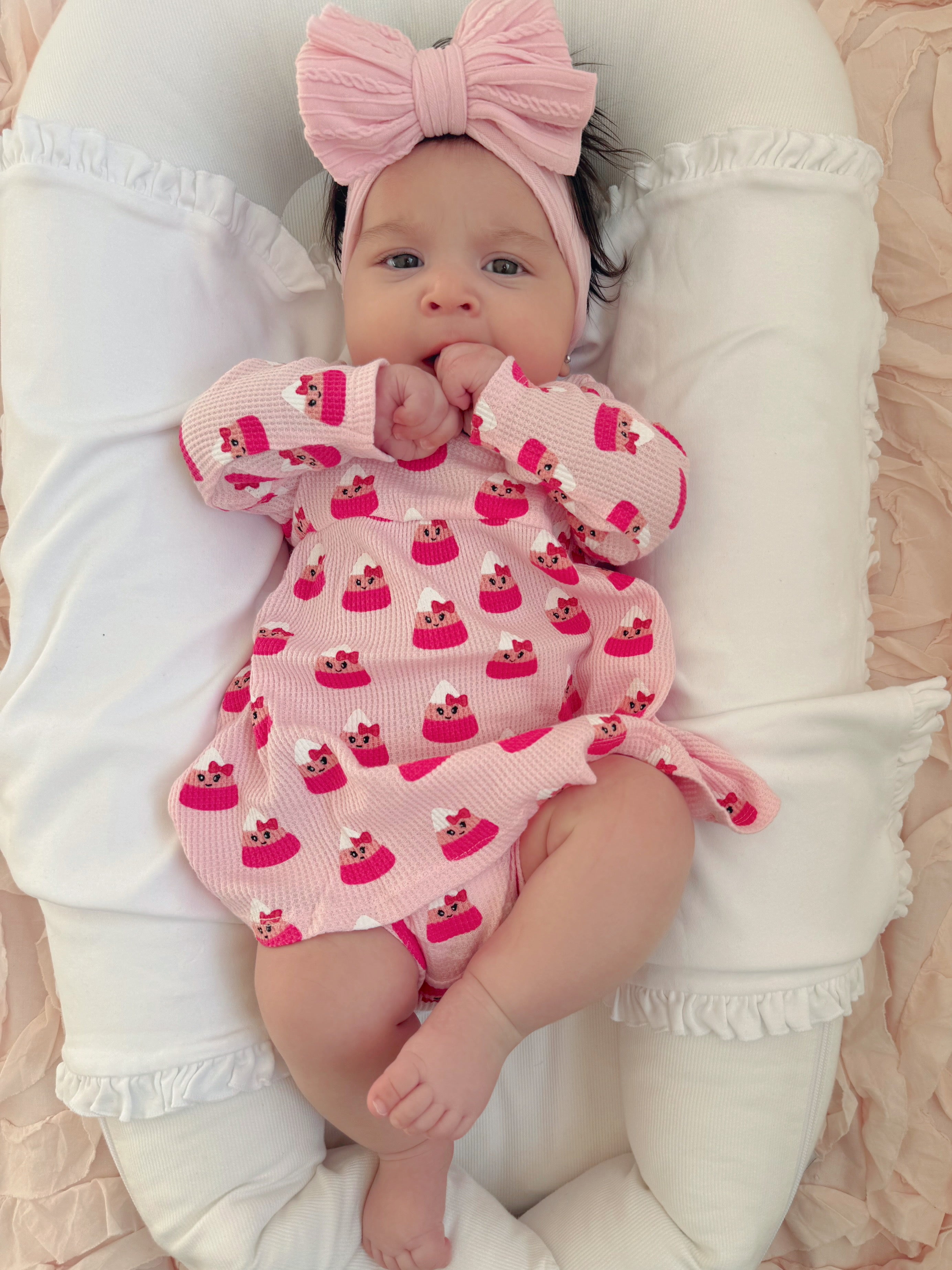 Smiling baby in a pink cupcake-patterned outfit, resting on a soft cushion with a large bow headband.
