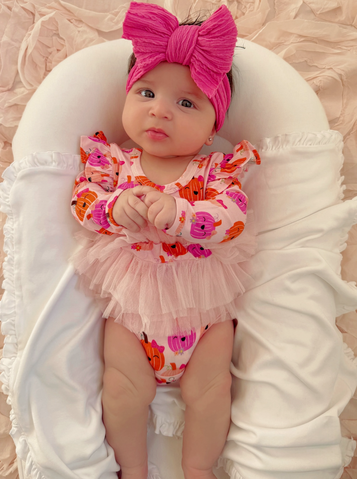 Baby girl in pink outfit with pumpkin print, wearing a large pink bow, sitting on a white plush surface.