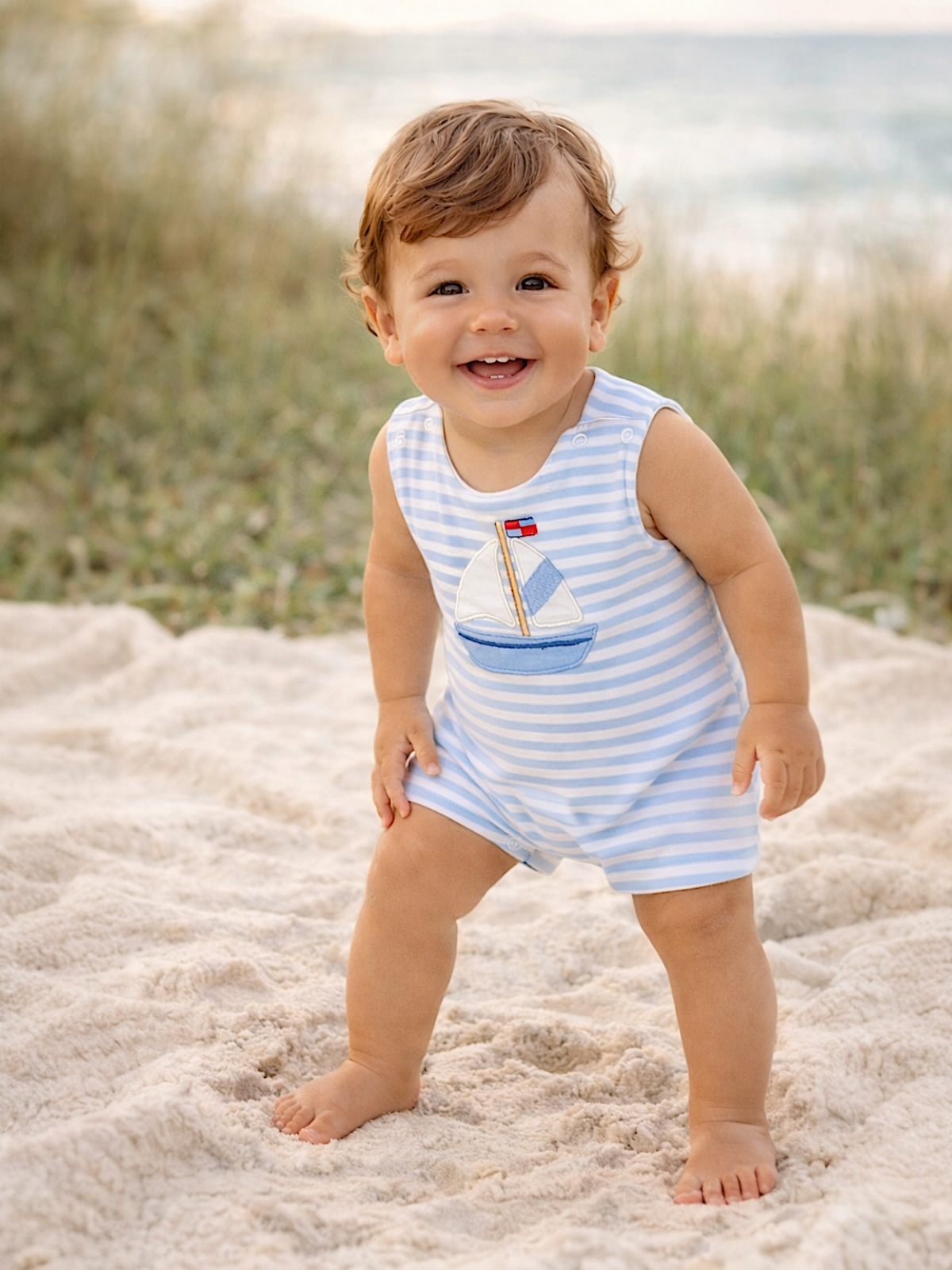 Smiling baby in blue striped romper stands on sandy beach, with grass and water in the background.