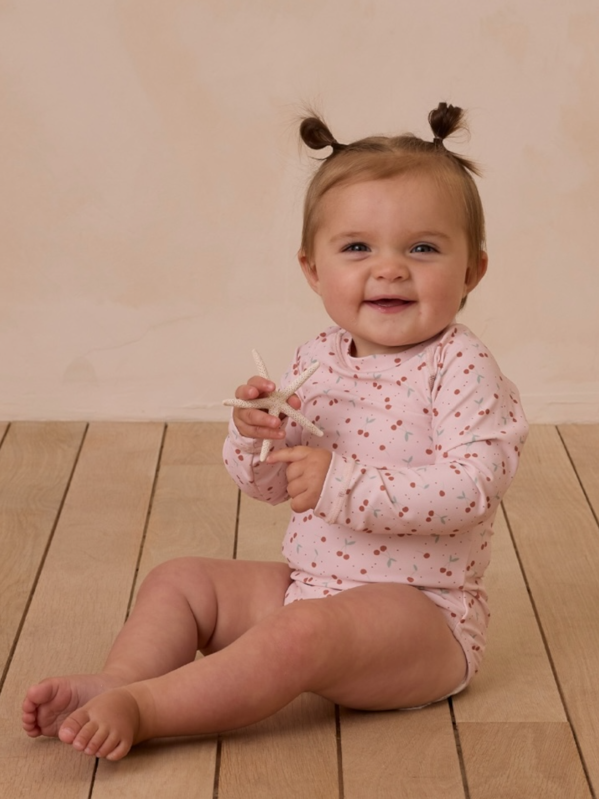 Smiling baby with pigtails wearing a pink outfit, sitting on wooden floor and holding a toy starfish.