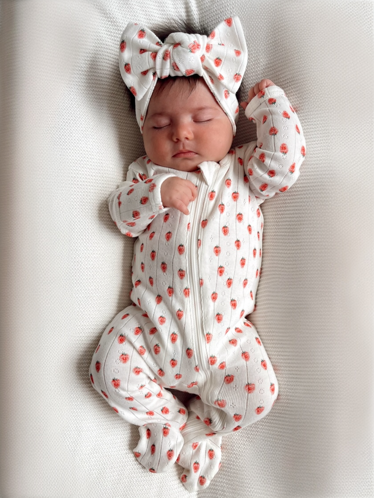 Infant sleeping in a strawberry-patterned onesie and matching bow headband, on a soft, light-colored background.