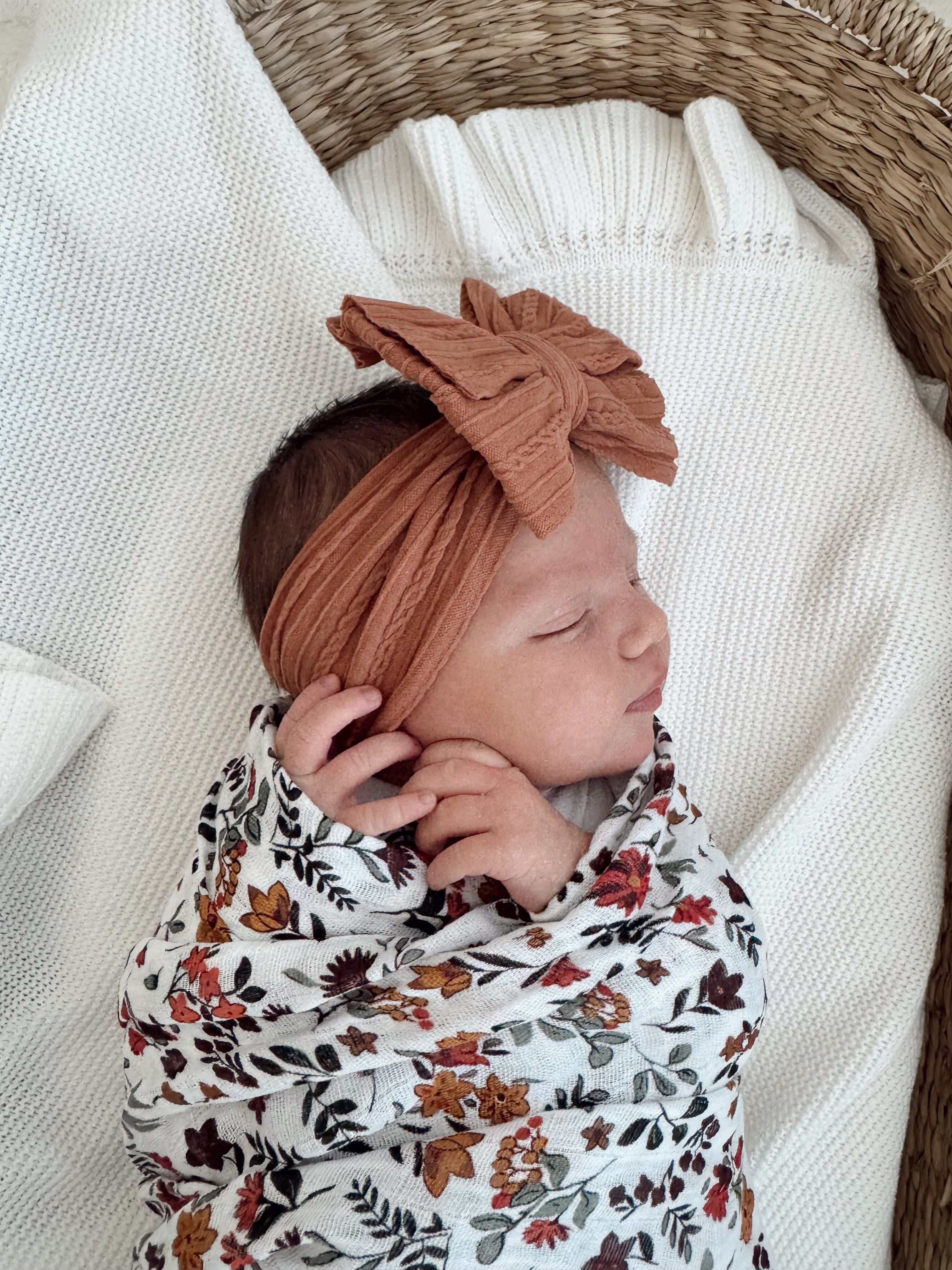 Newborn baby girl sleeping, wrapped in floral blanket with a rust-colored headband, resting on soft textures.