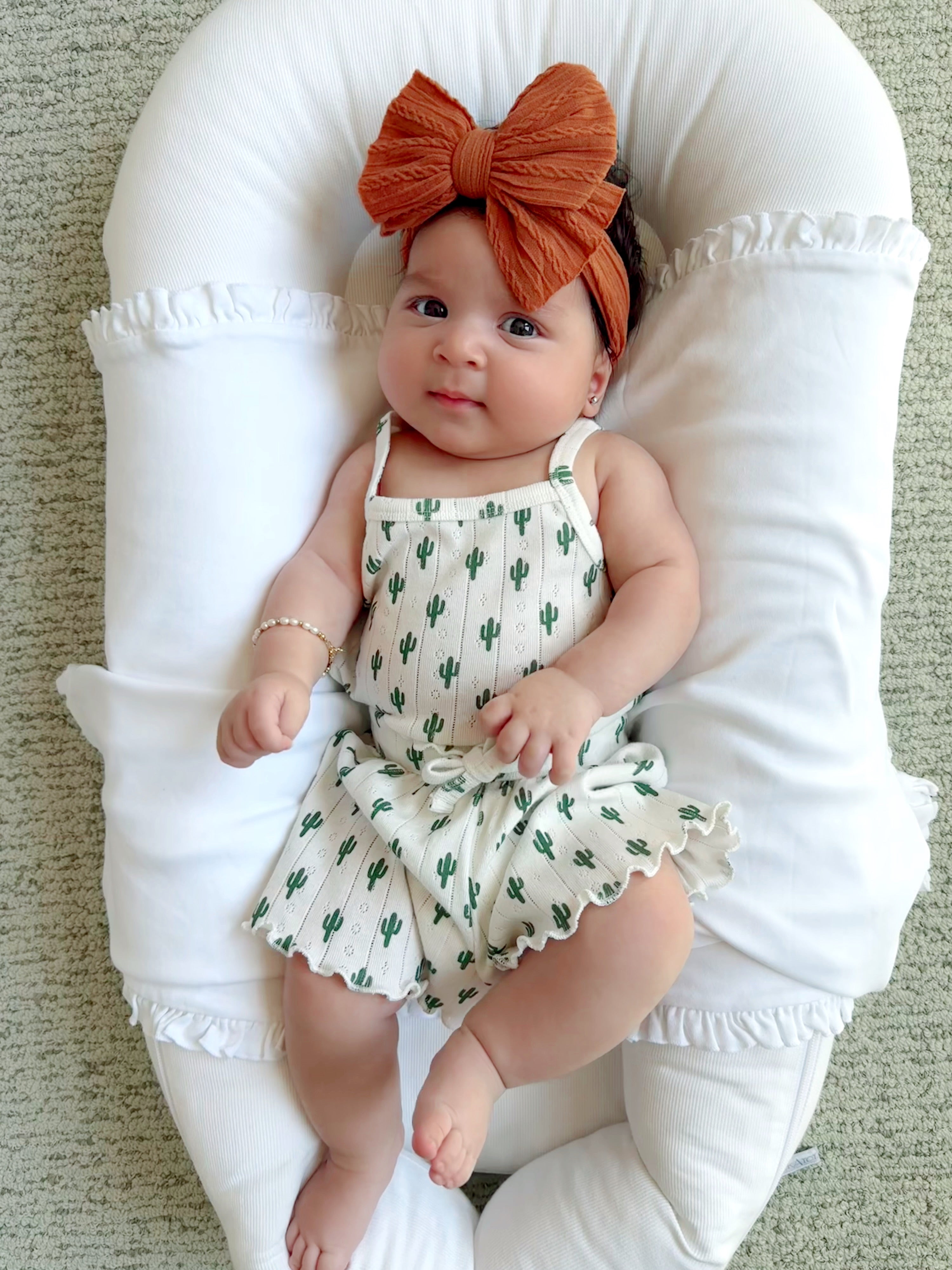 Infant in cactus-patterned romper, orange bow headband, lying on a white pillow on soft green carpet.