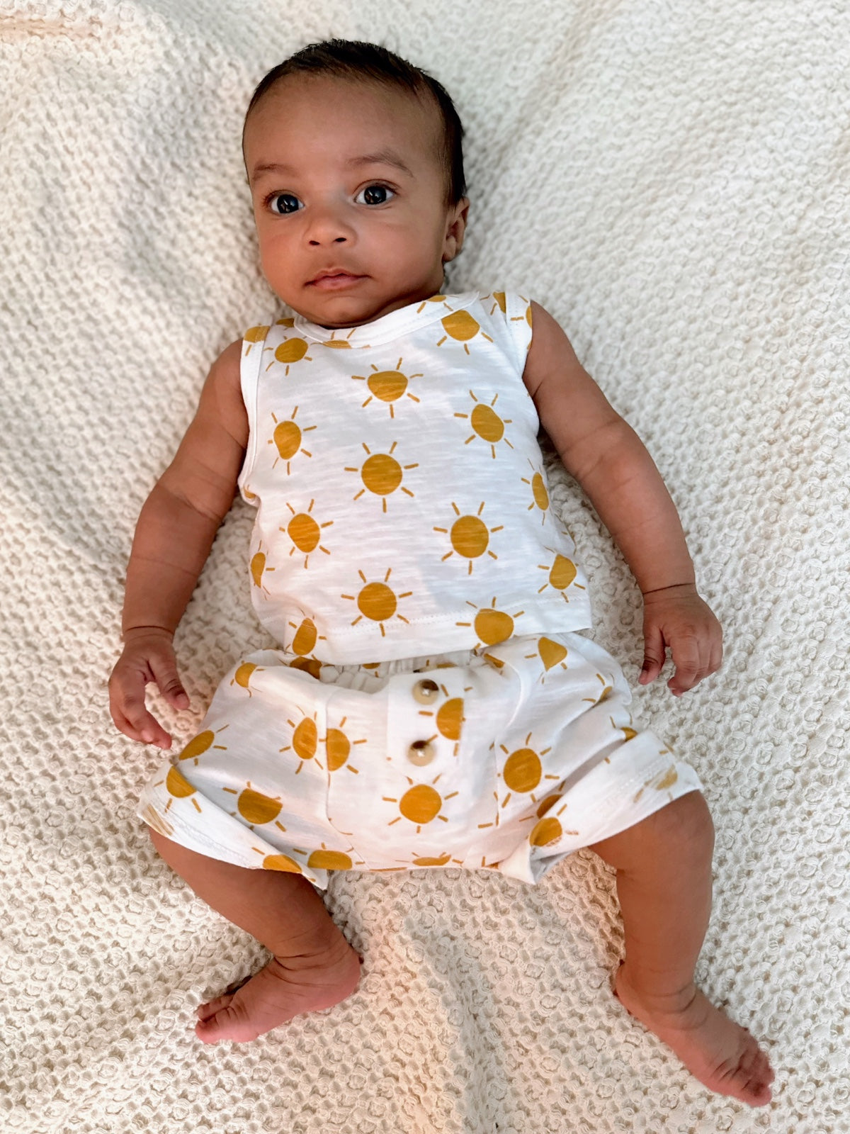 Infant lying on a fluffy blanket, wearing a sun-patterned outfit, with big expressive eyes.
