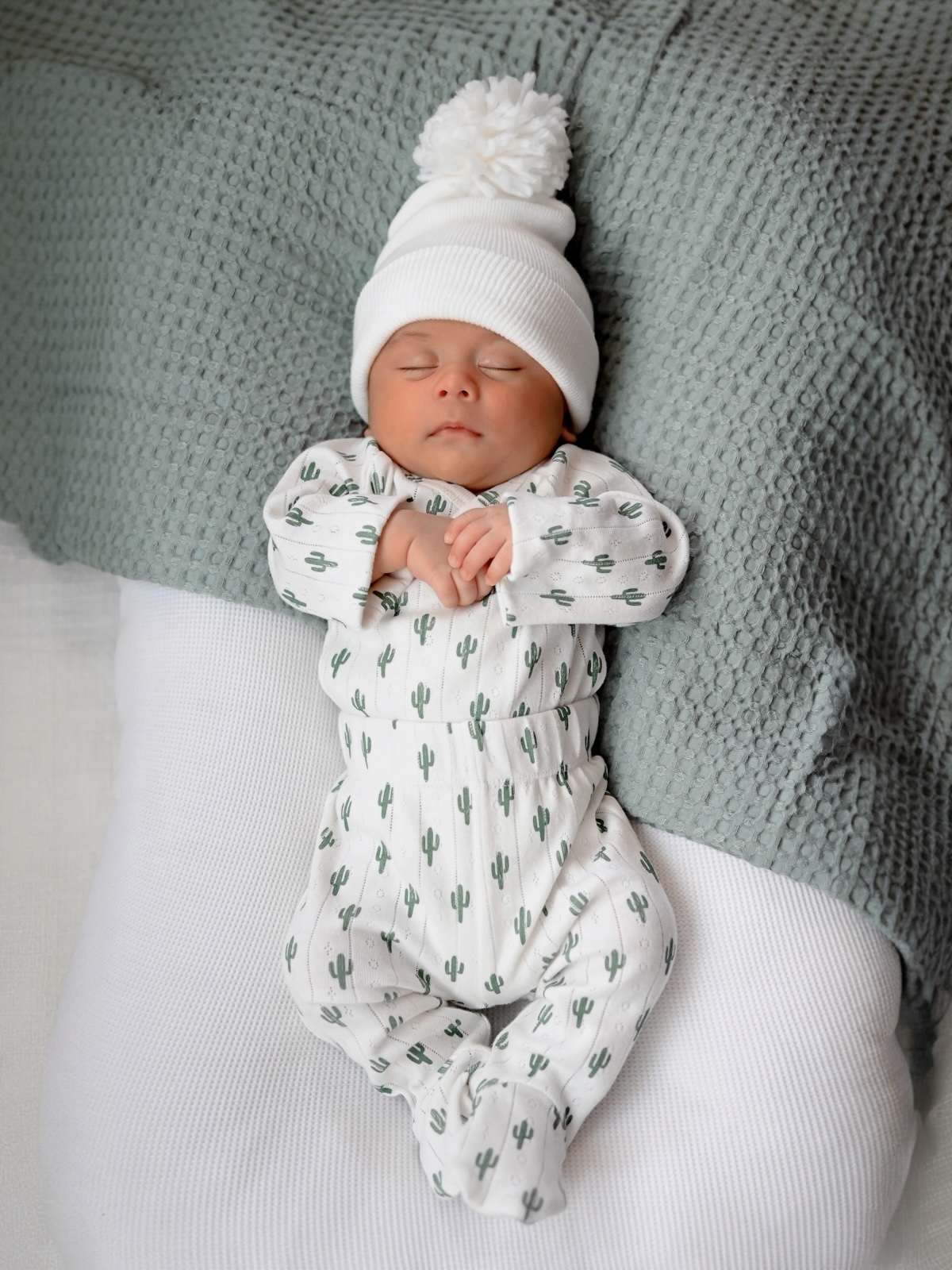 Newborn baby sleeping peacefully in a cactus-patterned outfit, wearing a white knit hat with a pom-pom.
