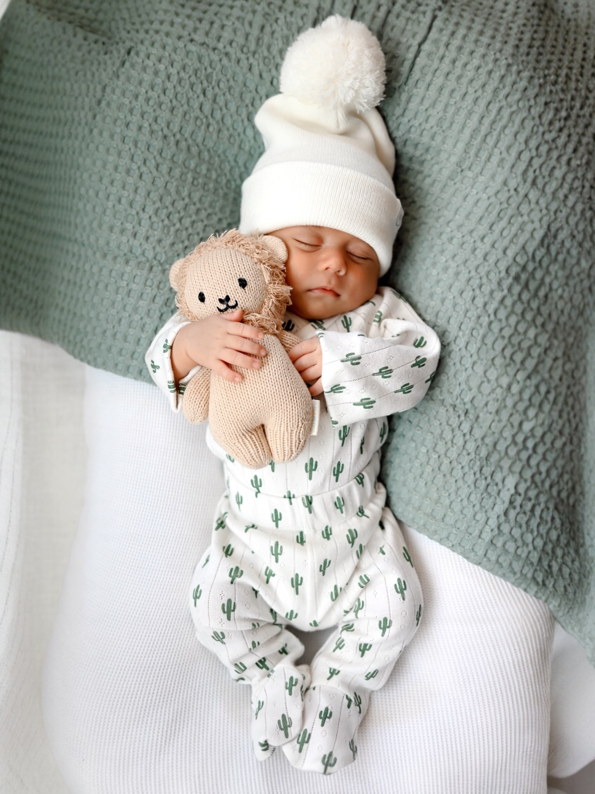 Baby in cactus-patterned pajamas sleeps peacefully holding a soft lion toy, cozy under a textured blanket.