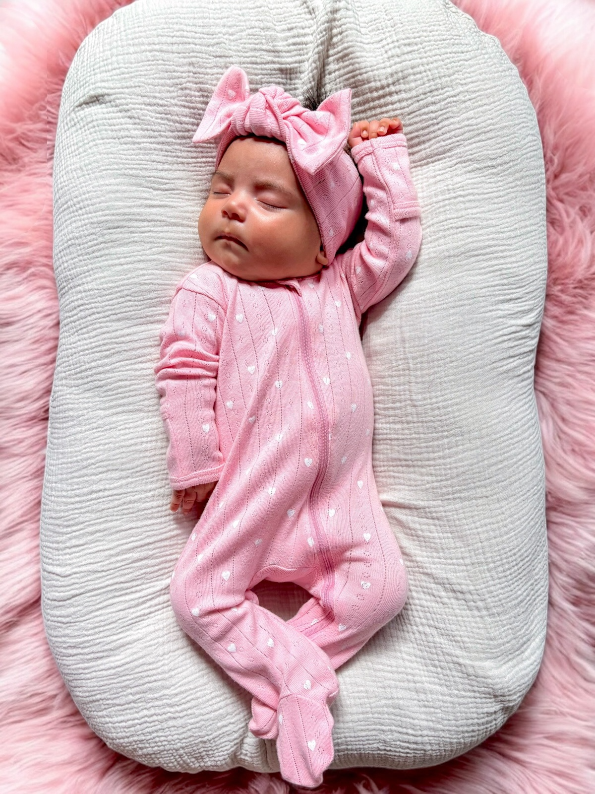 Baby girl in a pink heart-patterned onesie and matching headband, peacefully sleeping on a soft white cushion.
