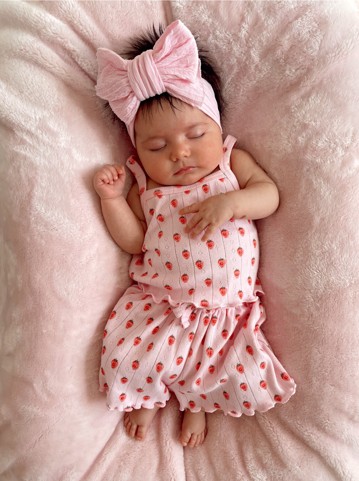 Infant girl asleep on pink blanket, wearing a pink strawberry-patterned outfit and large pink bow headband.