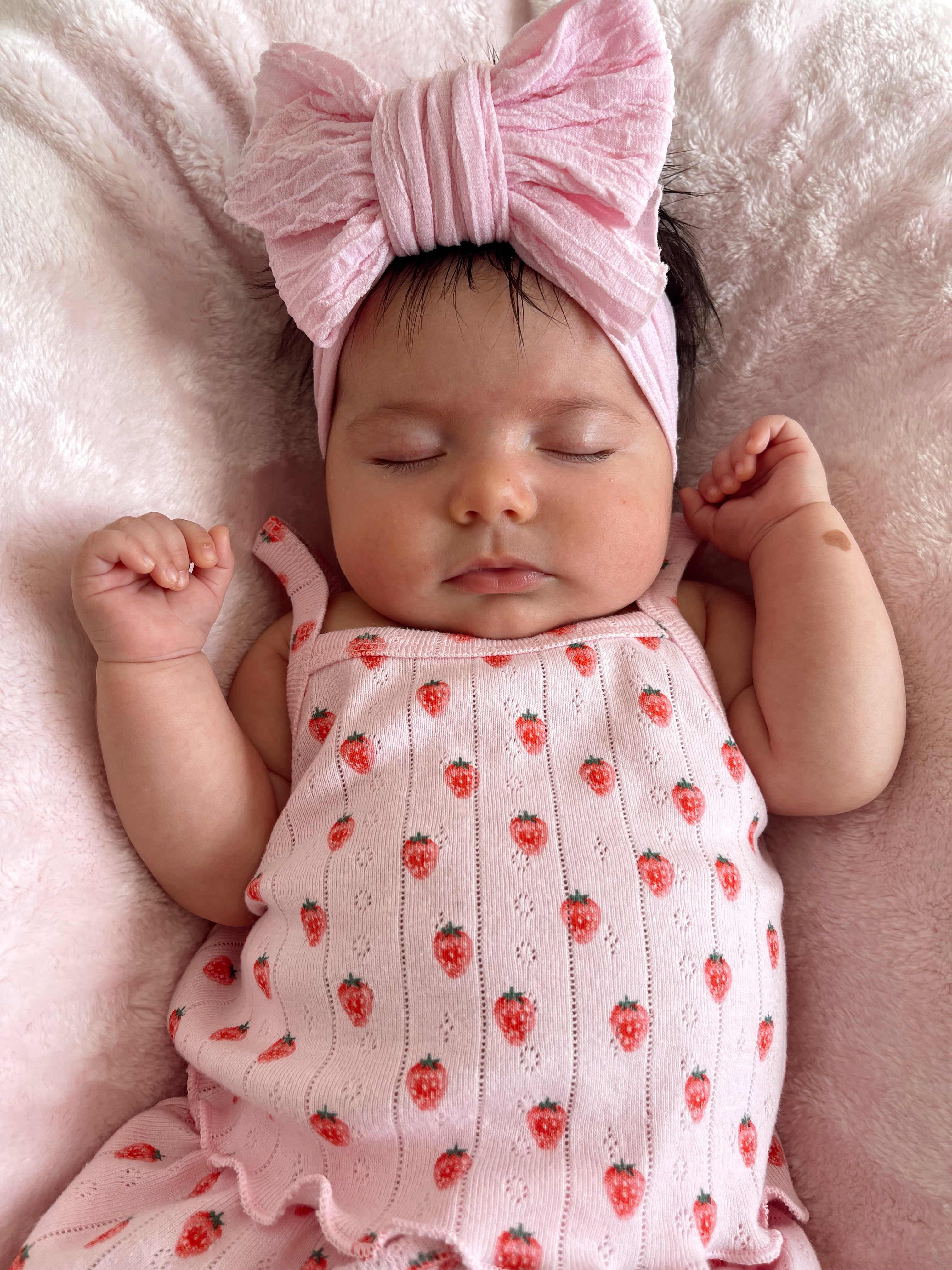 Sleeping baby girl in a pink outfit with strawberry patterns, wearing a large pink bow headband on a soft blanket.