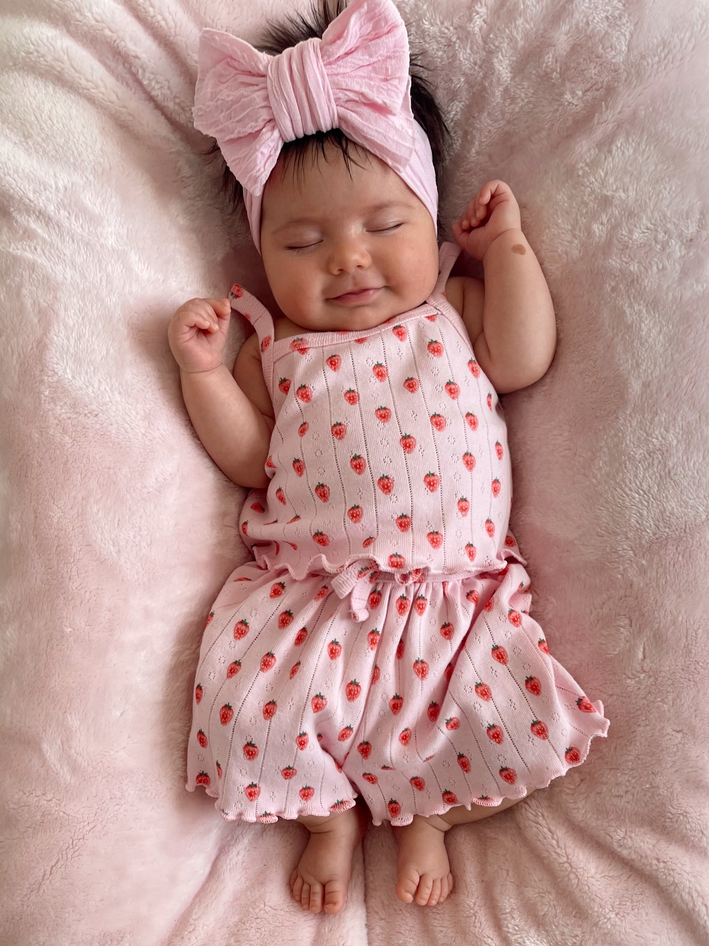Smiling baby in a pink strawberry-patterned outfit with a large bow headband, lying on a soft pink blanket.