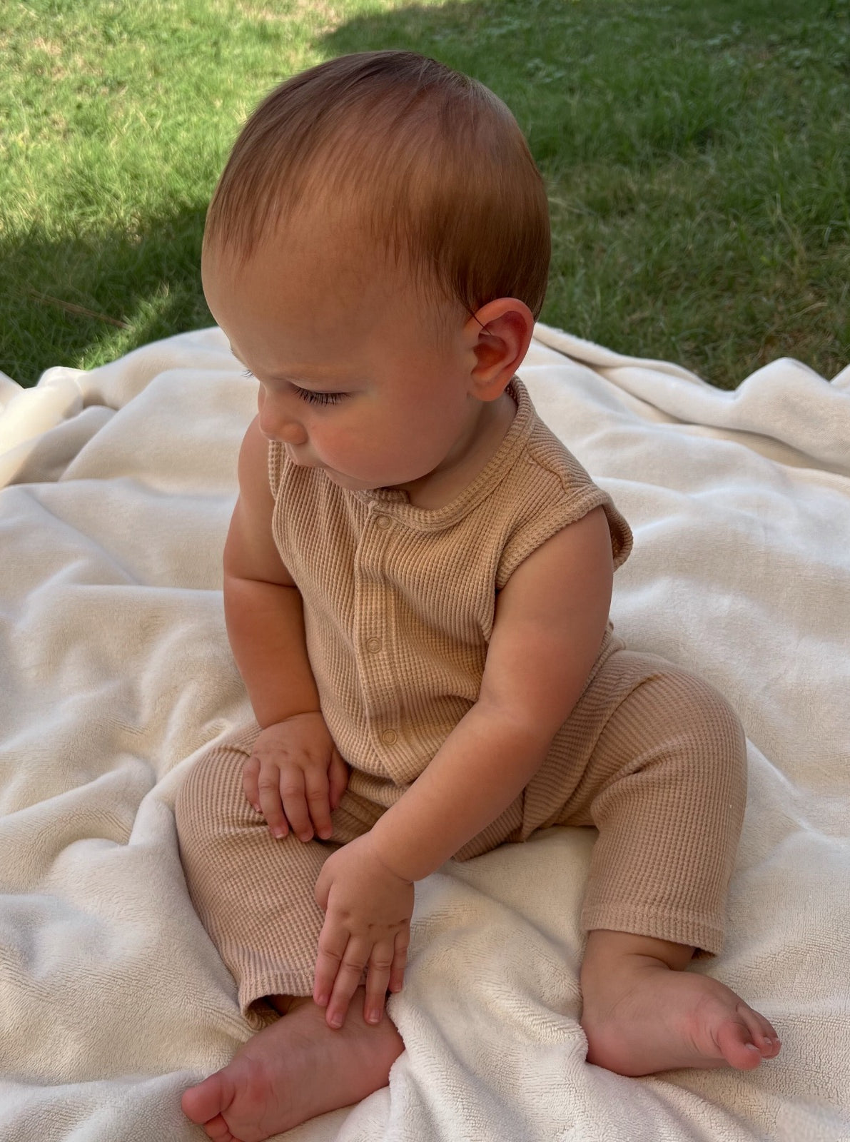 Baby sitting on a soft blanket outdoors, wearing a beige outfit, looking thoughtfully to the side.