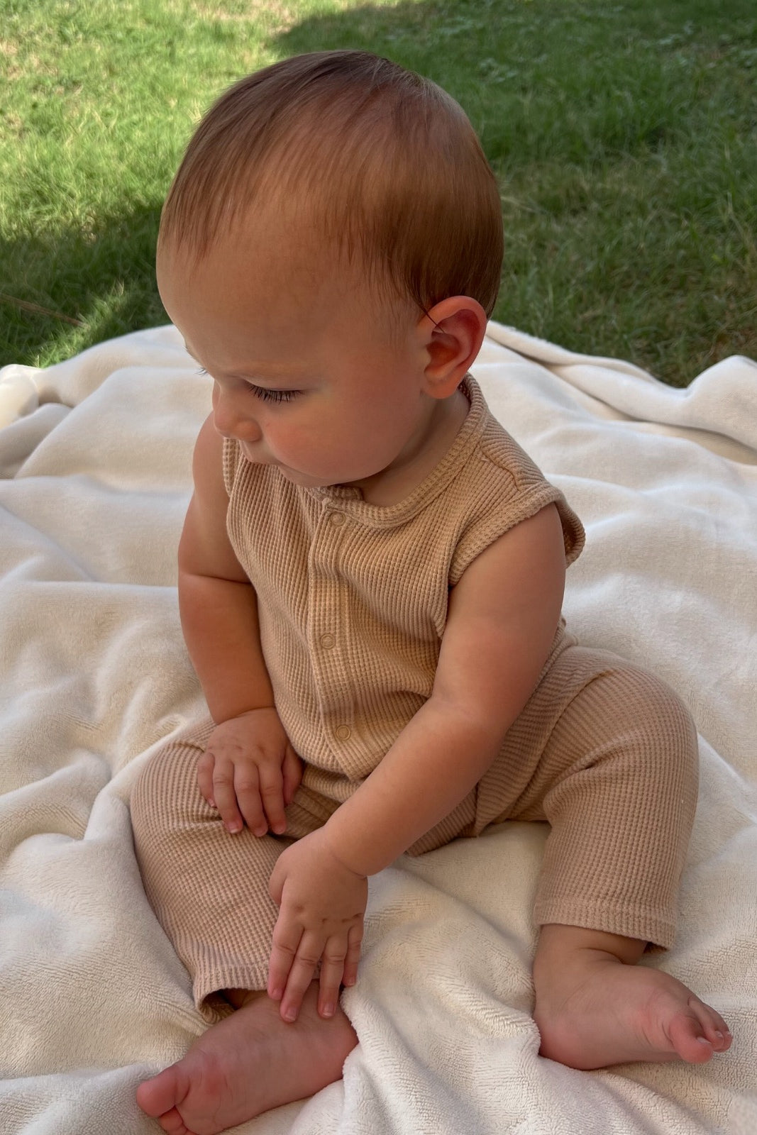 Baby sitting on a soft blanket outdoors, wearing a beige outfit, looking thoughtfully to the side.