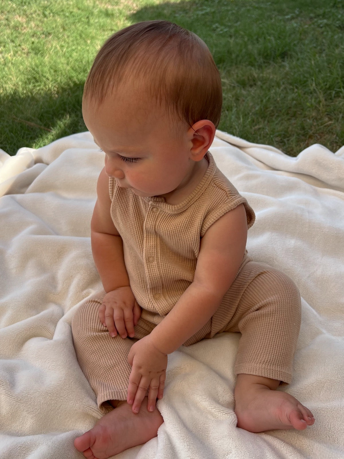 Baby sitting on a soft blanket outdoors, wearing a beige outfit, looking thoughtfully to the side.