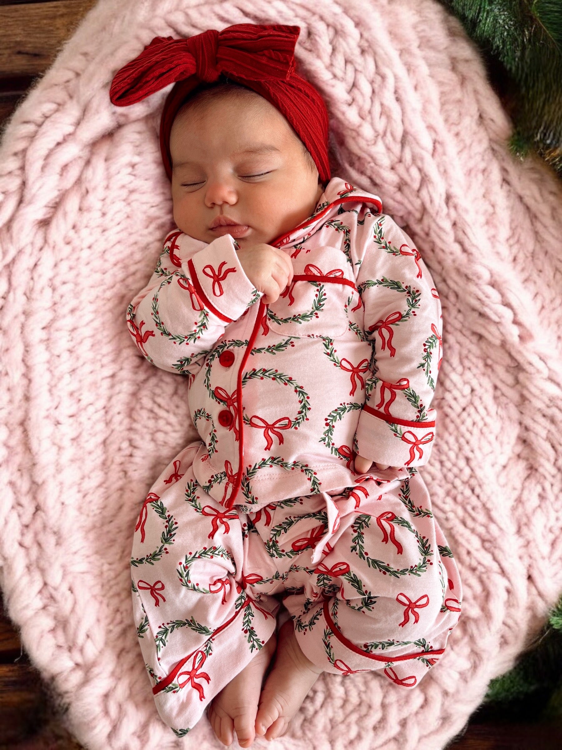 Baby girl in a pink holiday onesie with wreaths, resting on a soft pink blanket, wearing a red headband.