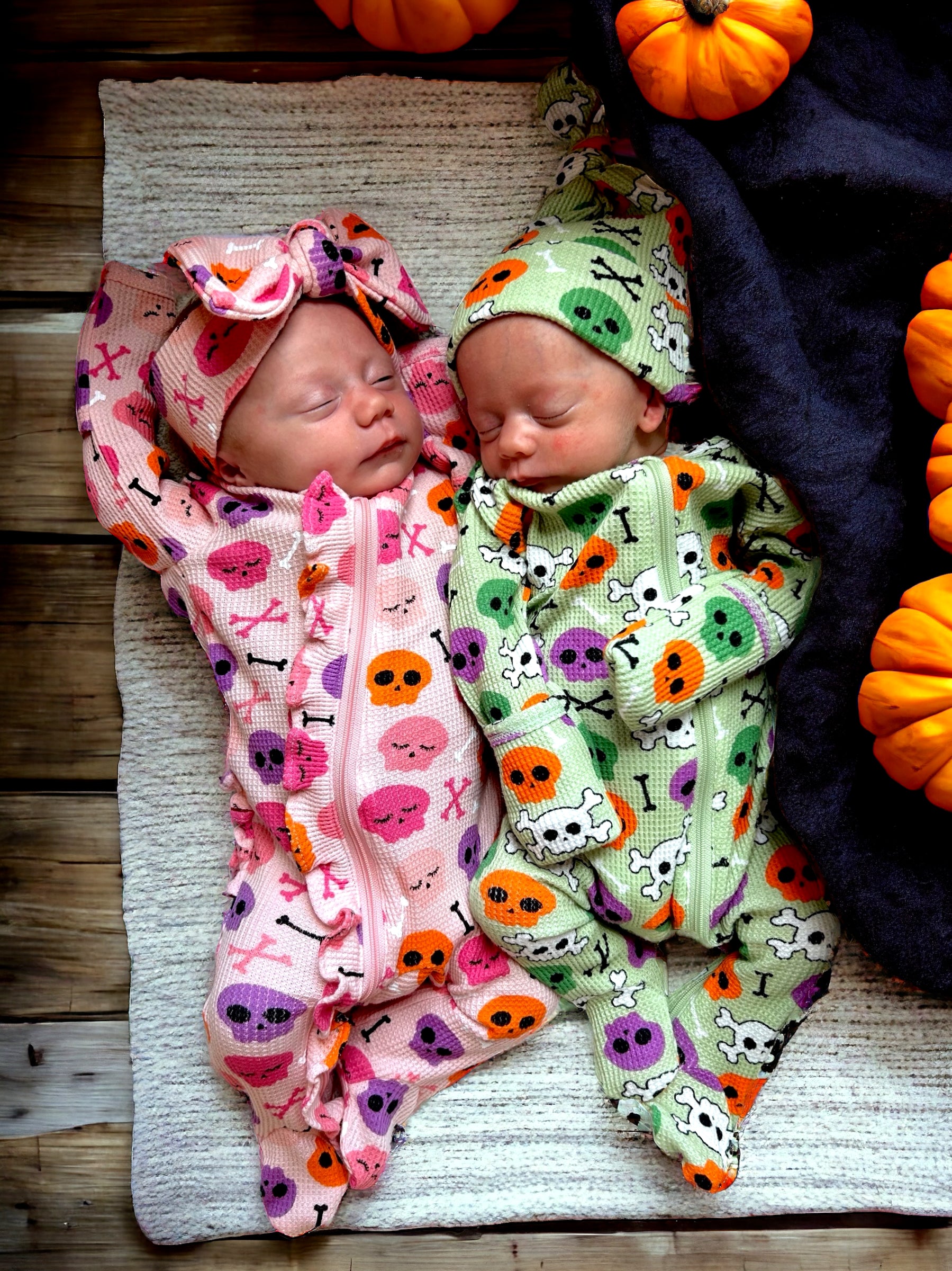Two newborn twins sleeping side by side in colorful themed outfits, surrounded by small pumpkins on a textured blanket.