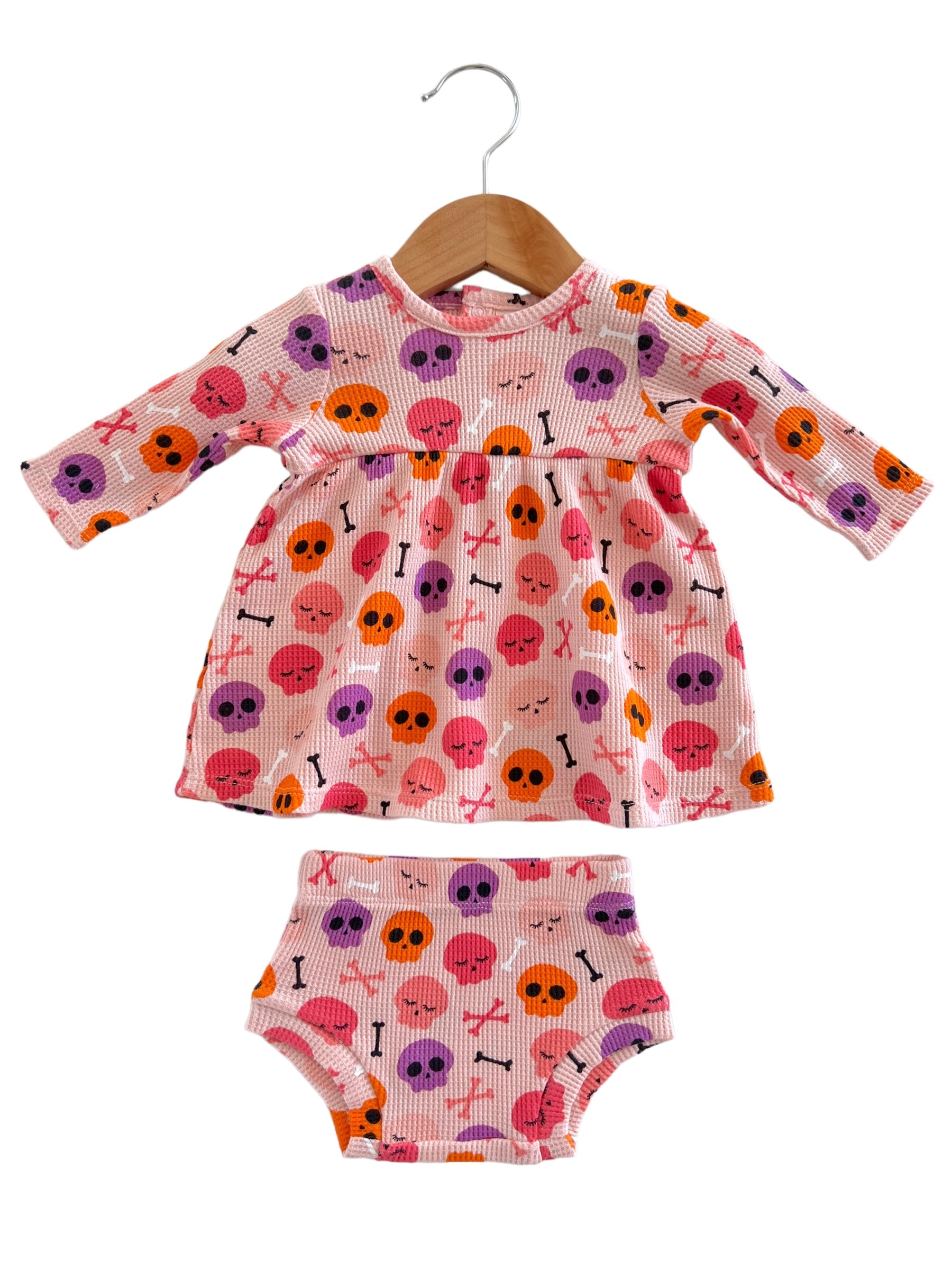 Pink dress and bloomers featuring colorful skull patterns, hanging on a wooden hanger. Perfect for kids' costumes.