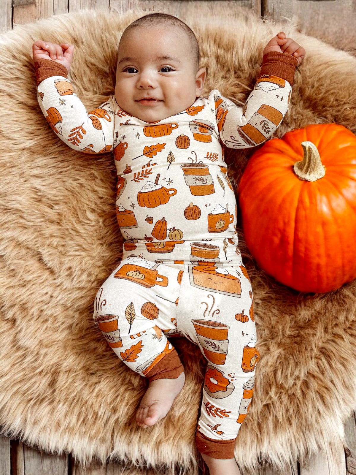 Smiling baby in cozy autumn-themed outfit next to a bright orange pumpkin, lying on soft fur.
