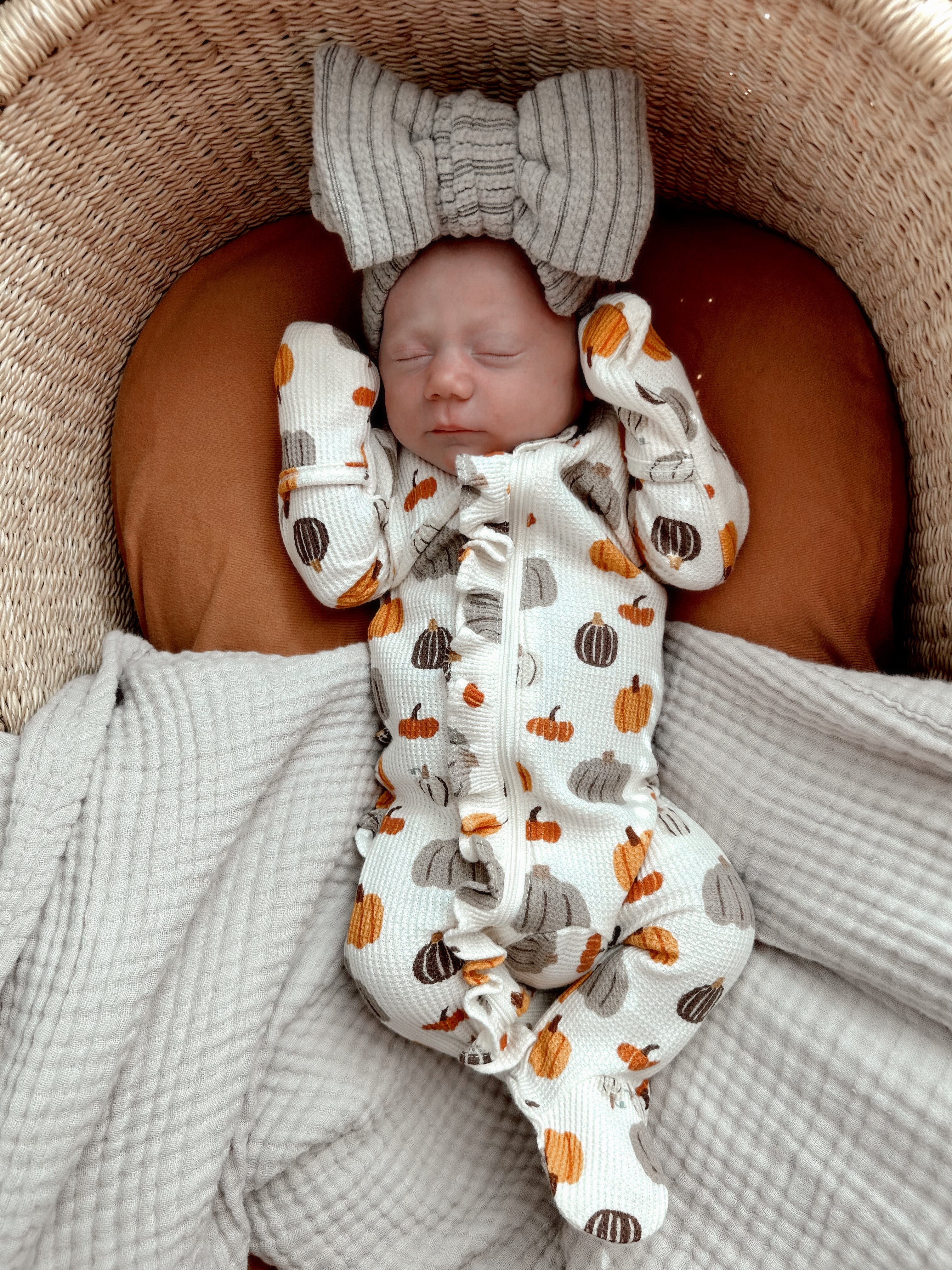 Newborn baby peacefully sleeping in a pumpkin-patterned outfit and large bow headband on a cozy blanket.