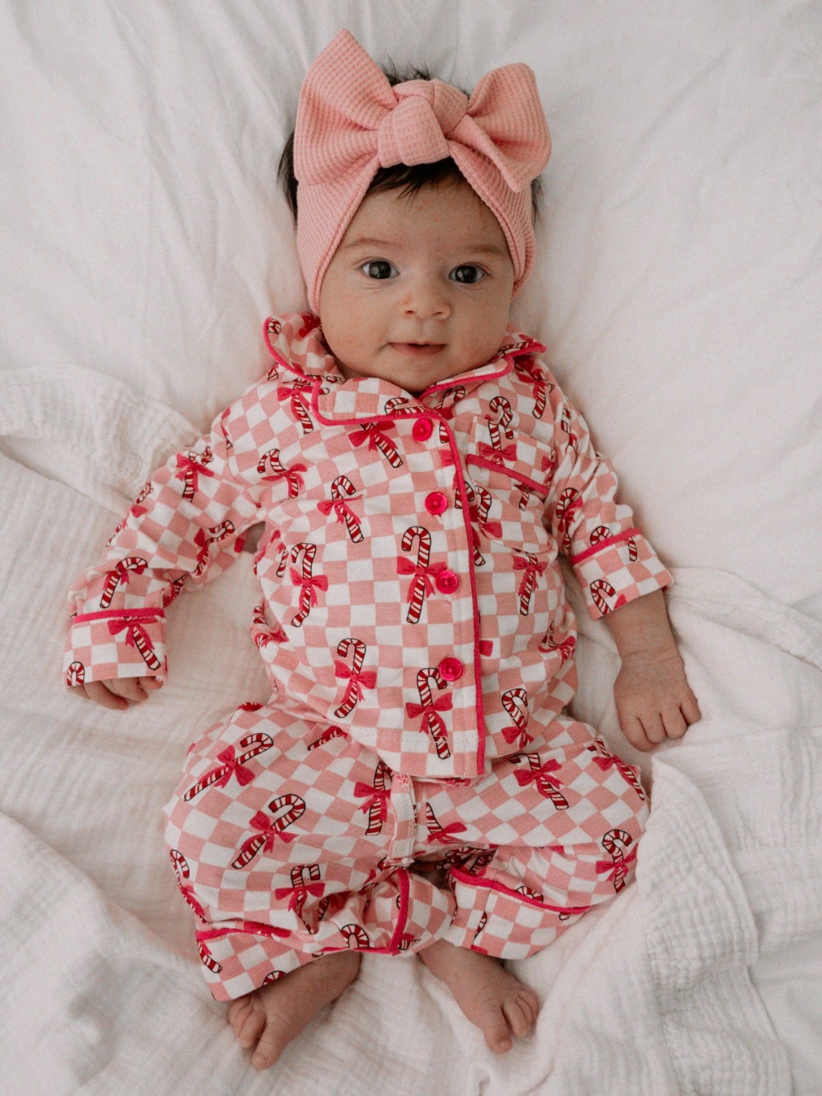Baby in candy-cane patterned pajamas and a pink bow headband, lying on a soft blanket.