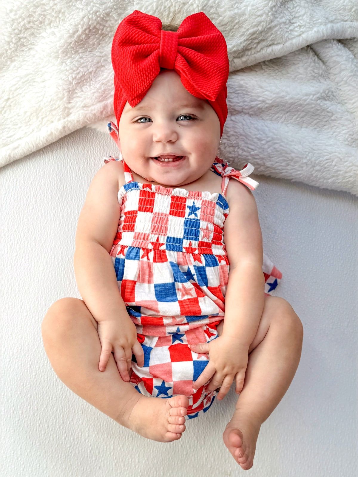 Smiling baby with blue eyes wearing a red bow and a checkered outfit, sitting on a soft white surface.