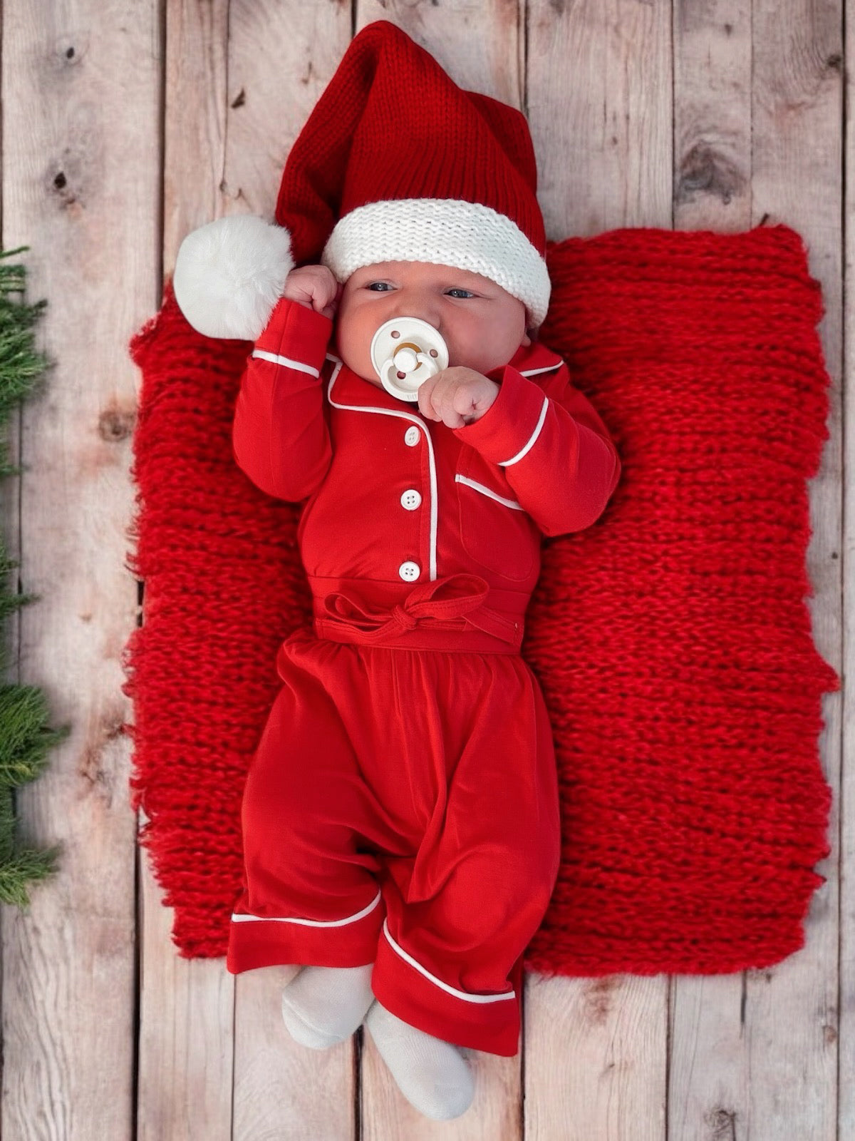 Baby in a festive red outfit and Santa hat, resting on a red blanket, against a wooden backdrop.
