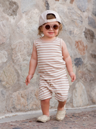 Smiling toddler in striped outfit, cap, and sunglasses, walking on a stone path.
