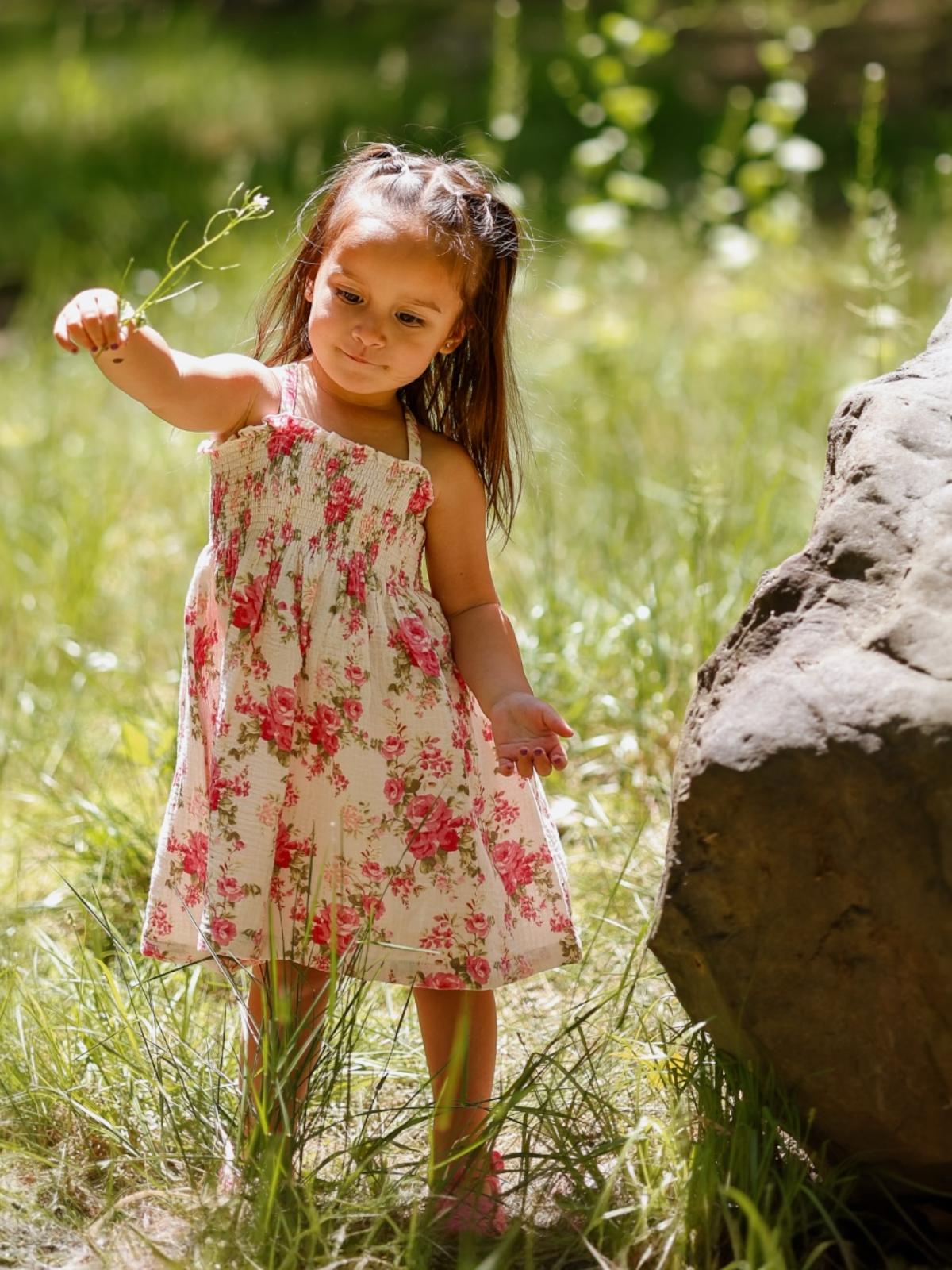 A young girl in a floral dress examines a small plant in a sunlit grassy area.
