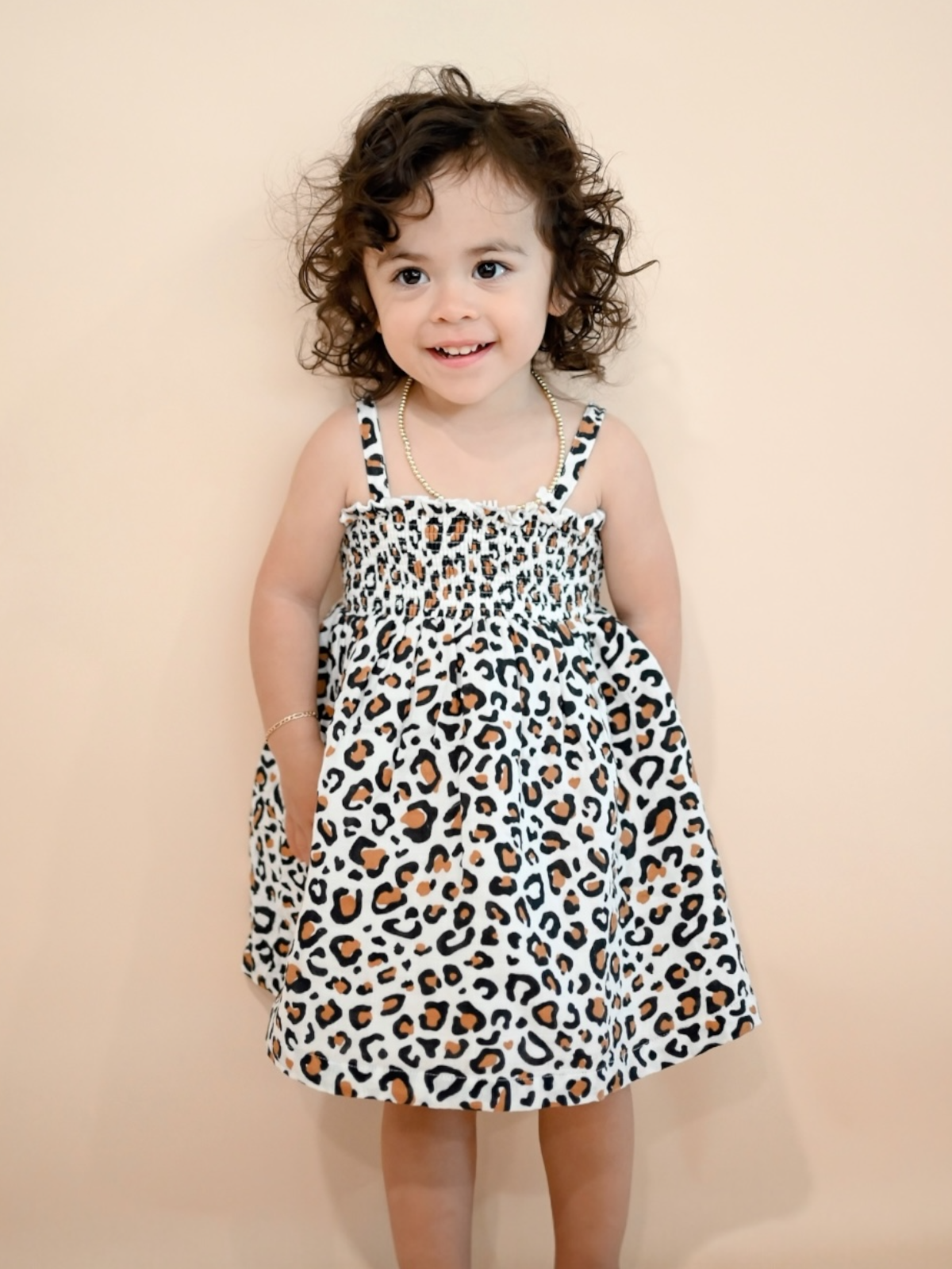 Smiling child with curly hair wearing a leopard-print dress, standing against a neutral background.