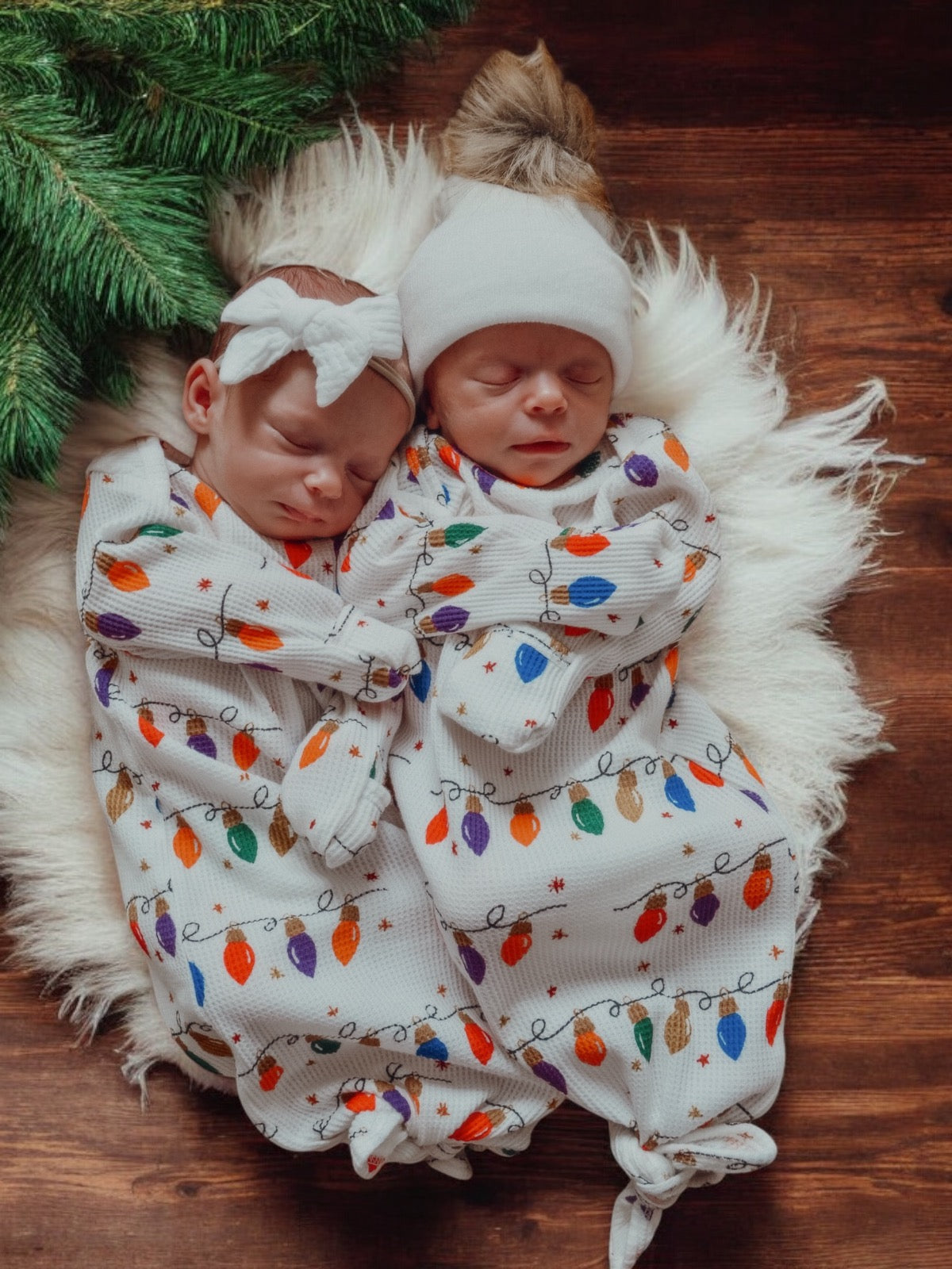 Two sleeping infants in festive onesies with holiday lights, lying on a cozy white blanket, surrounded by greenery.