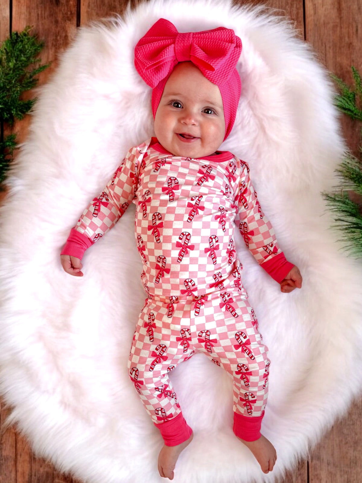 Smiling baby in pink candy cane outfit and bow, lying on fluffy white blanket with greenery in background.