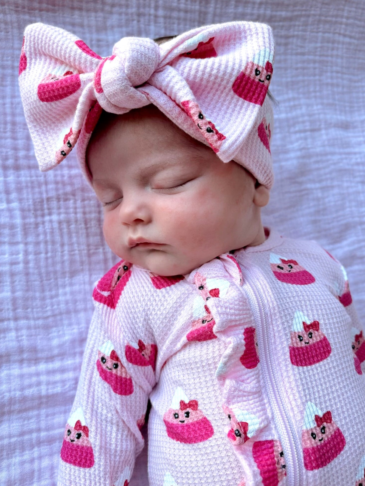 Baby girl sleeping in a pink cupcake-patterned outfit with a large bow headband on a textured blanket.