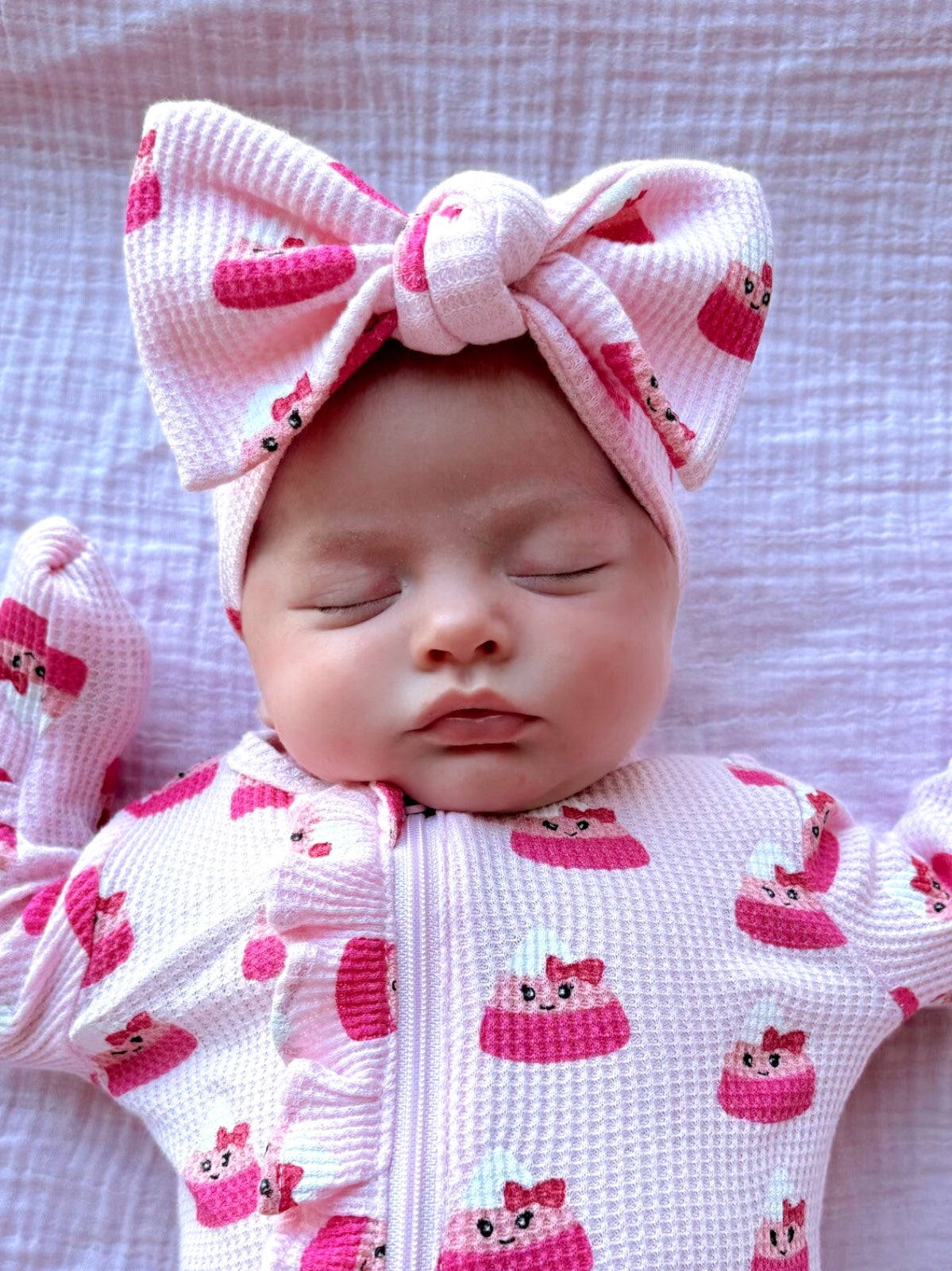 A sleeping baby in a pink outfit with cake patterns and a large bow headband. Soft, peaceful expression on a textured blanket.