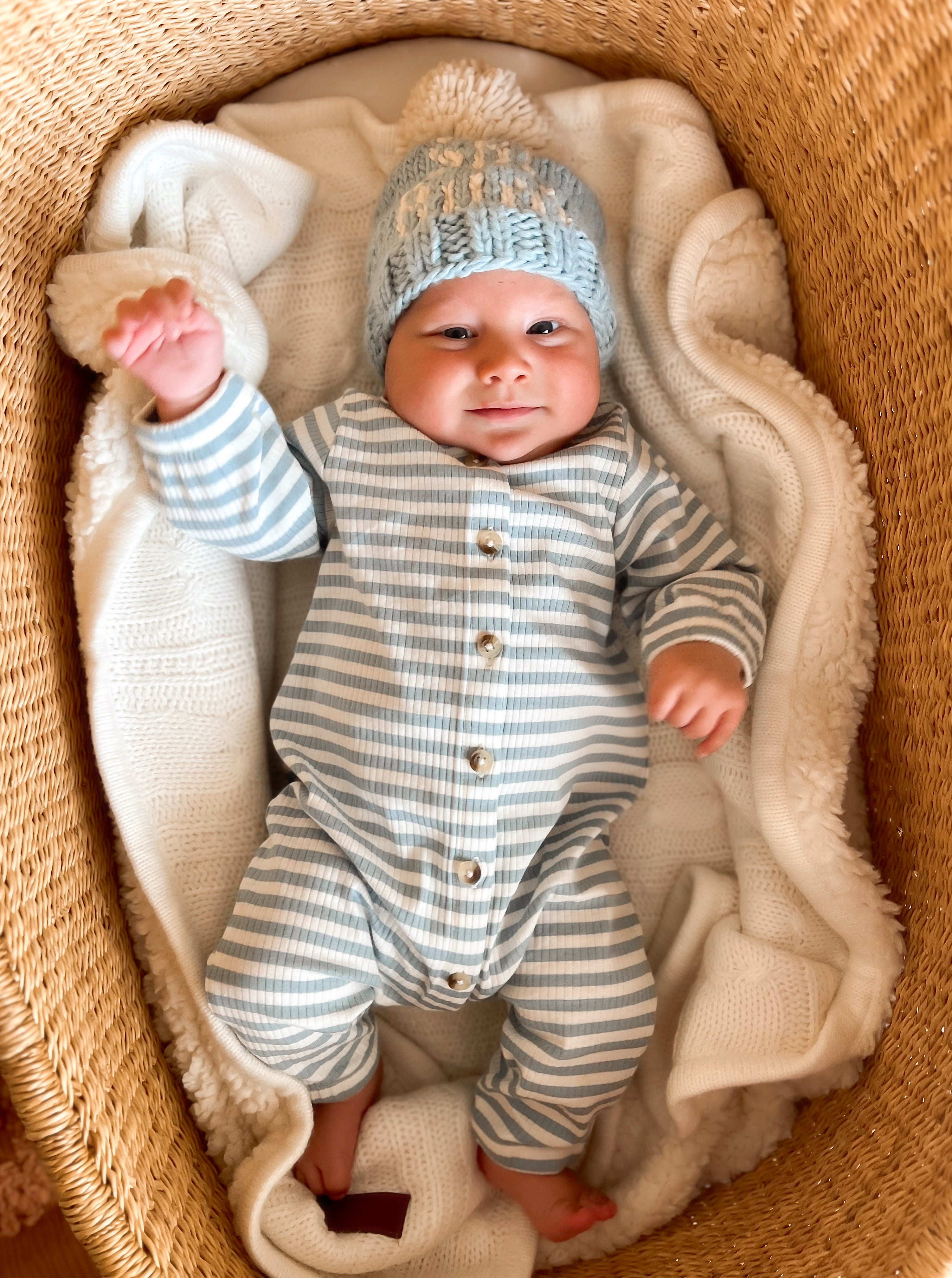 Smiling baby in a striped outfit and knitted hat, lying in a cozy basket surrounded by a blanket.