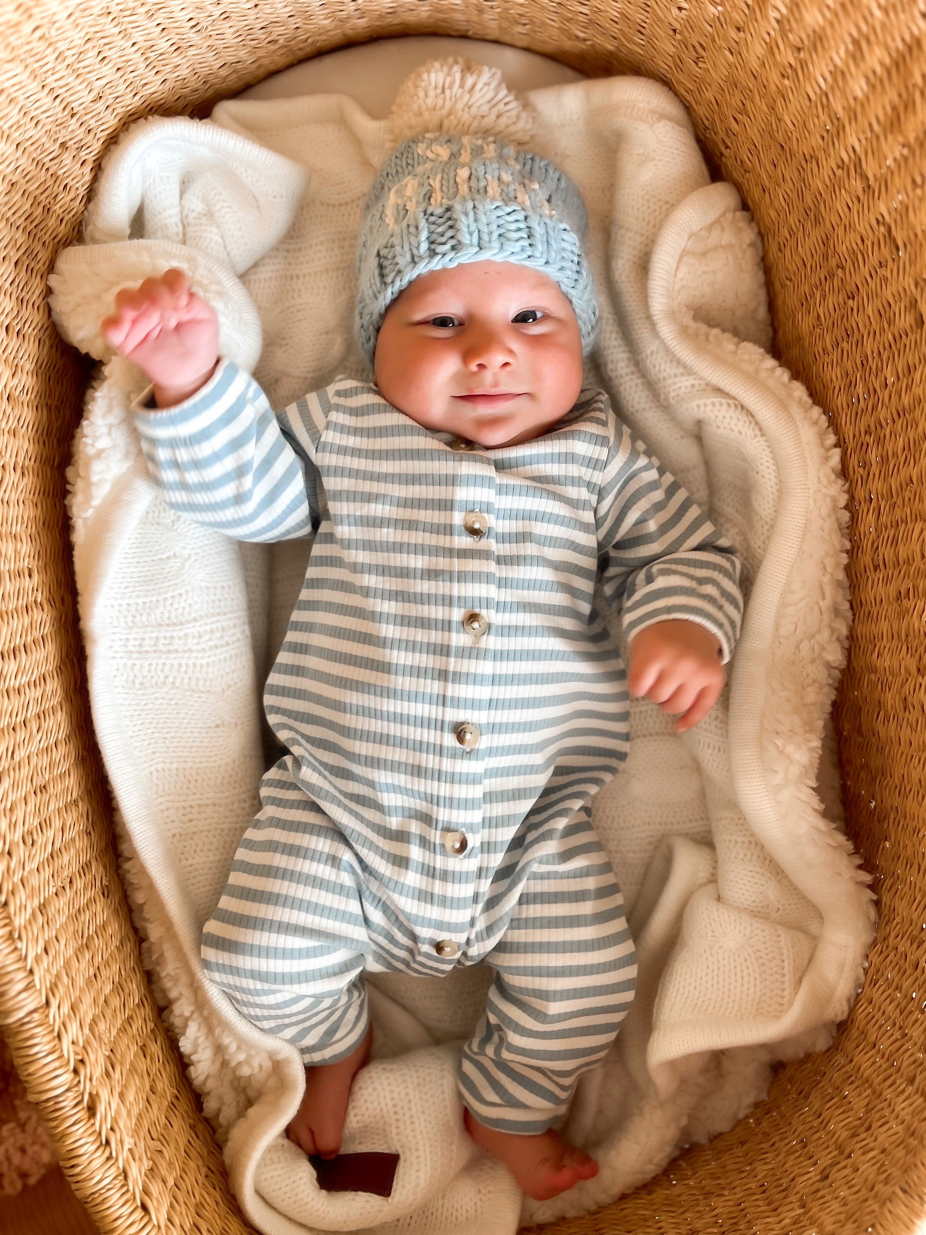 Smiling baby in a striped outfit and knitted hat, lying in a cozy basket surrounded by a blanket.
