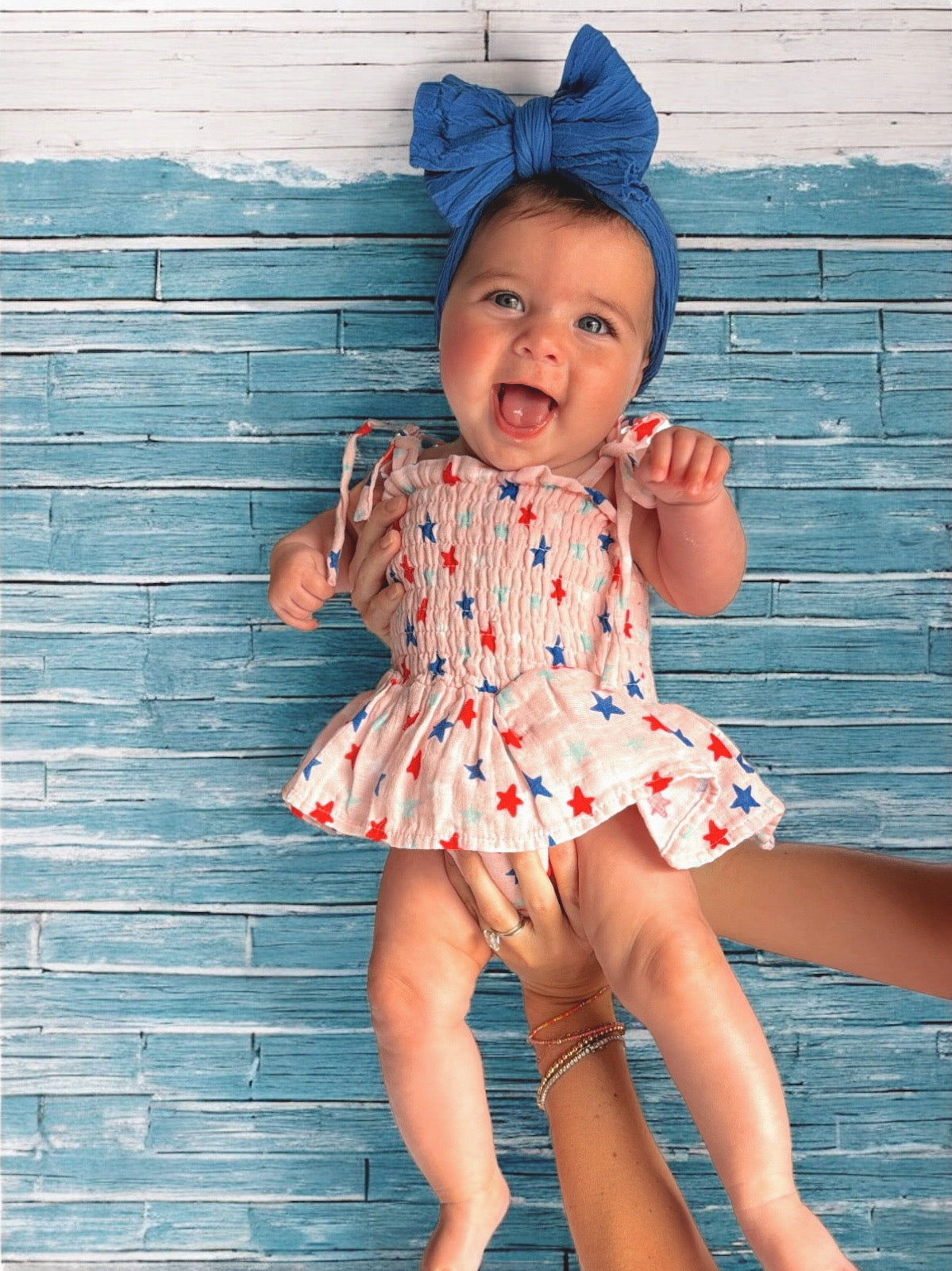 Smiling baby girl in a star-patterned dress, held against a blue wooden background, wearing a blue headband.