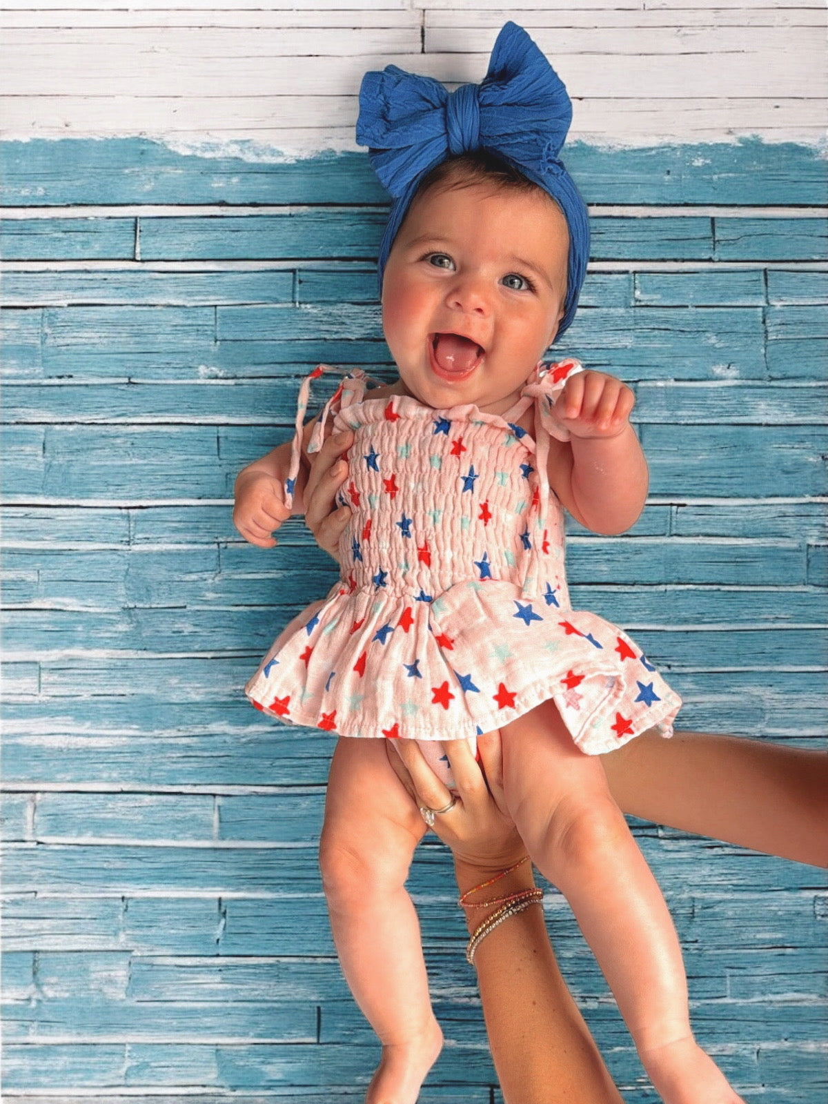 Smiling baby girl in a star-patterned dress, held against a blue wooden background, wearing a blue headband.