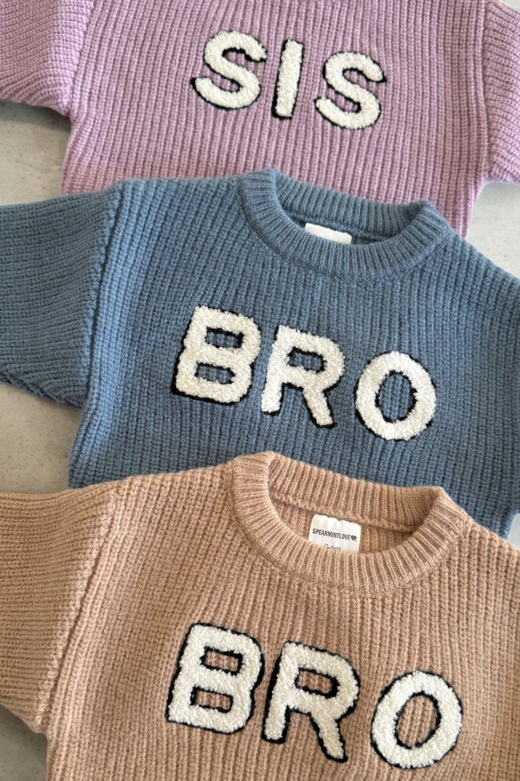 Three knit sweaters in pastel colors with the words "SIS" and "BRO" prominently displayed on them.