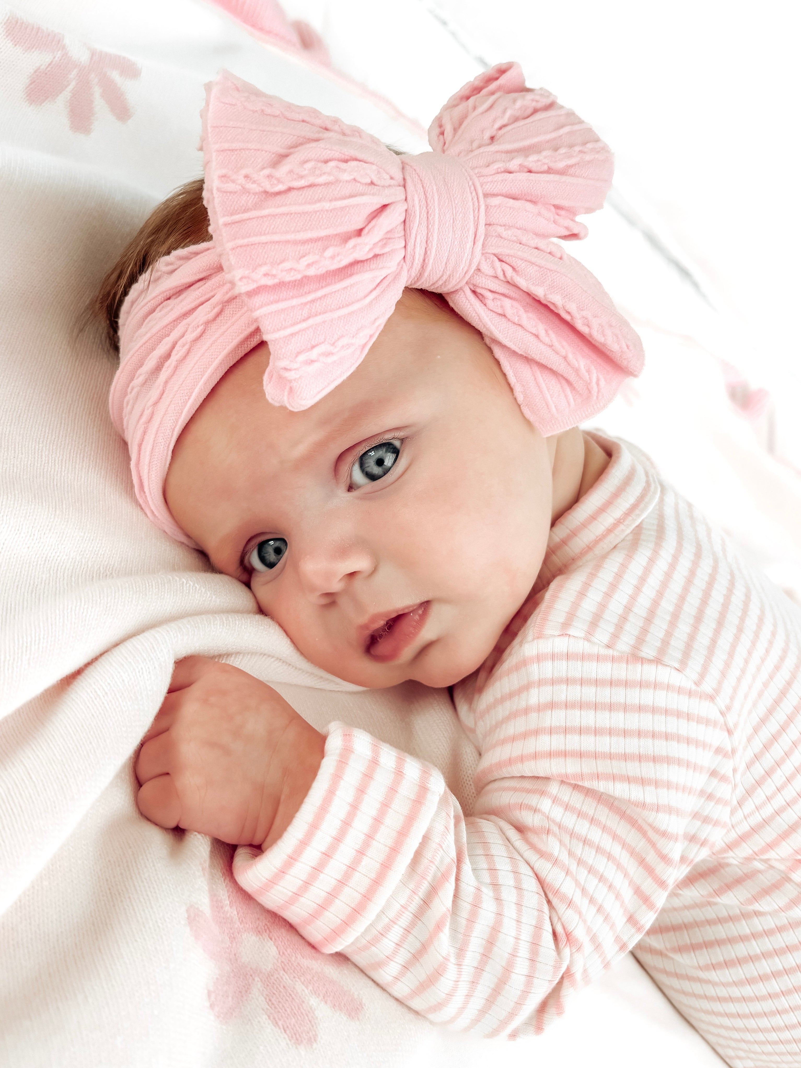 Baby with big pink headband, lying on a soft blanket, looking curiously at the camera.
