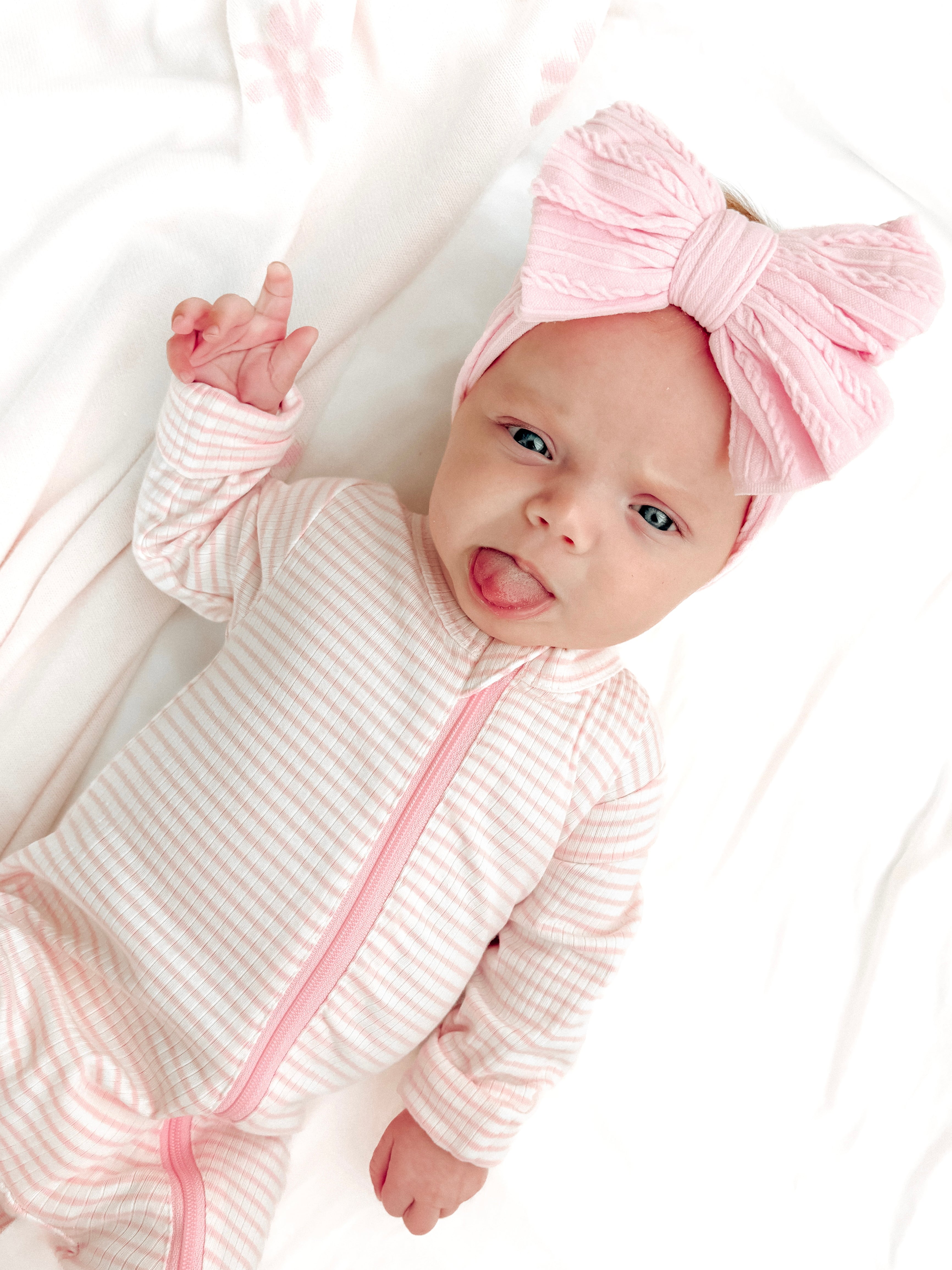 Baby in a striped onesie and large pink bow, playfully sticking out her tongue against a soft, light background.