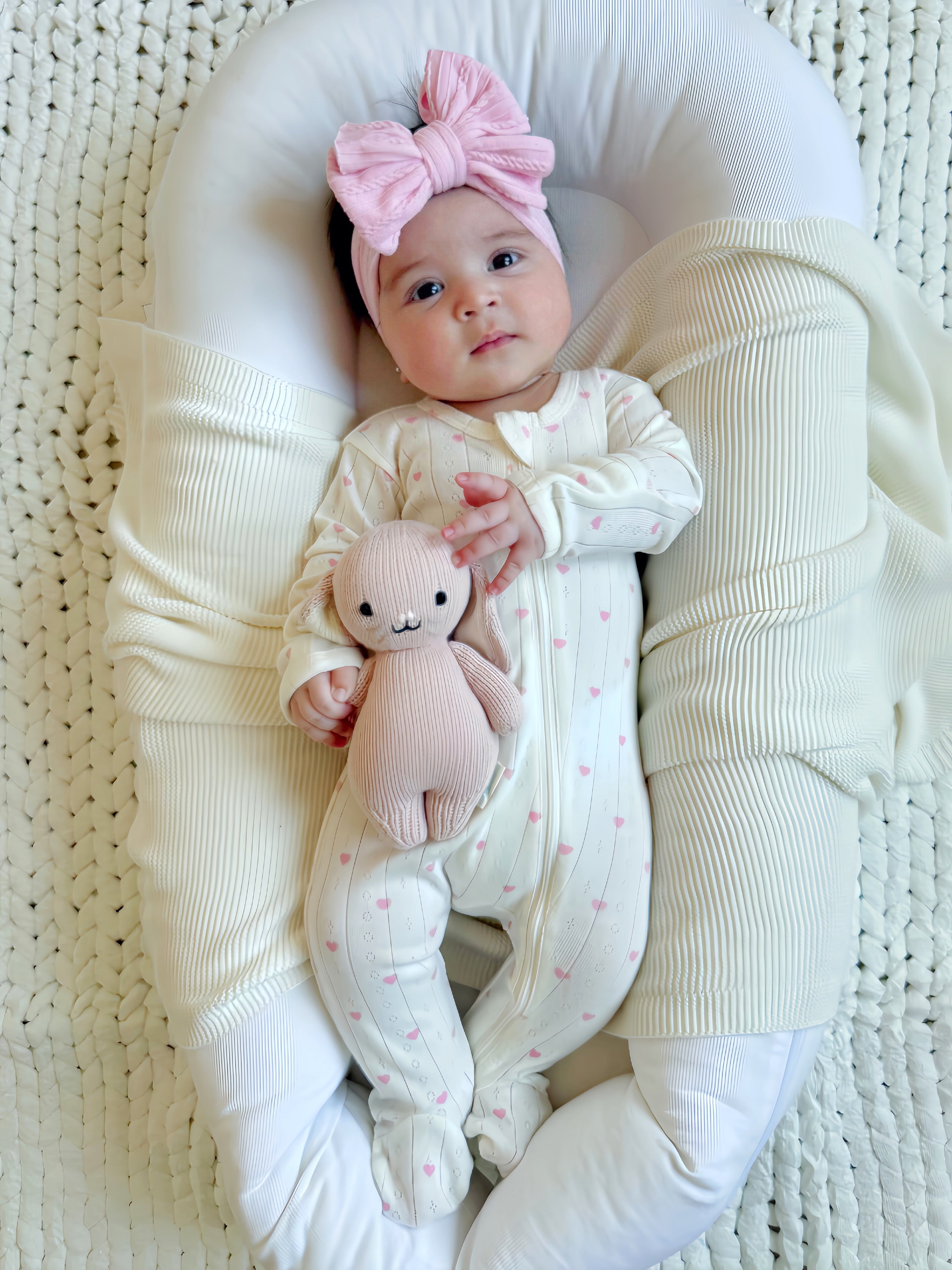 Baby in cozy crib wearing a patterned onesie, holding a soft toy, with a large pink bow in her hair.