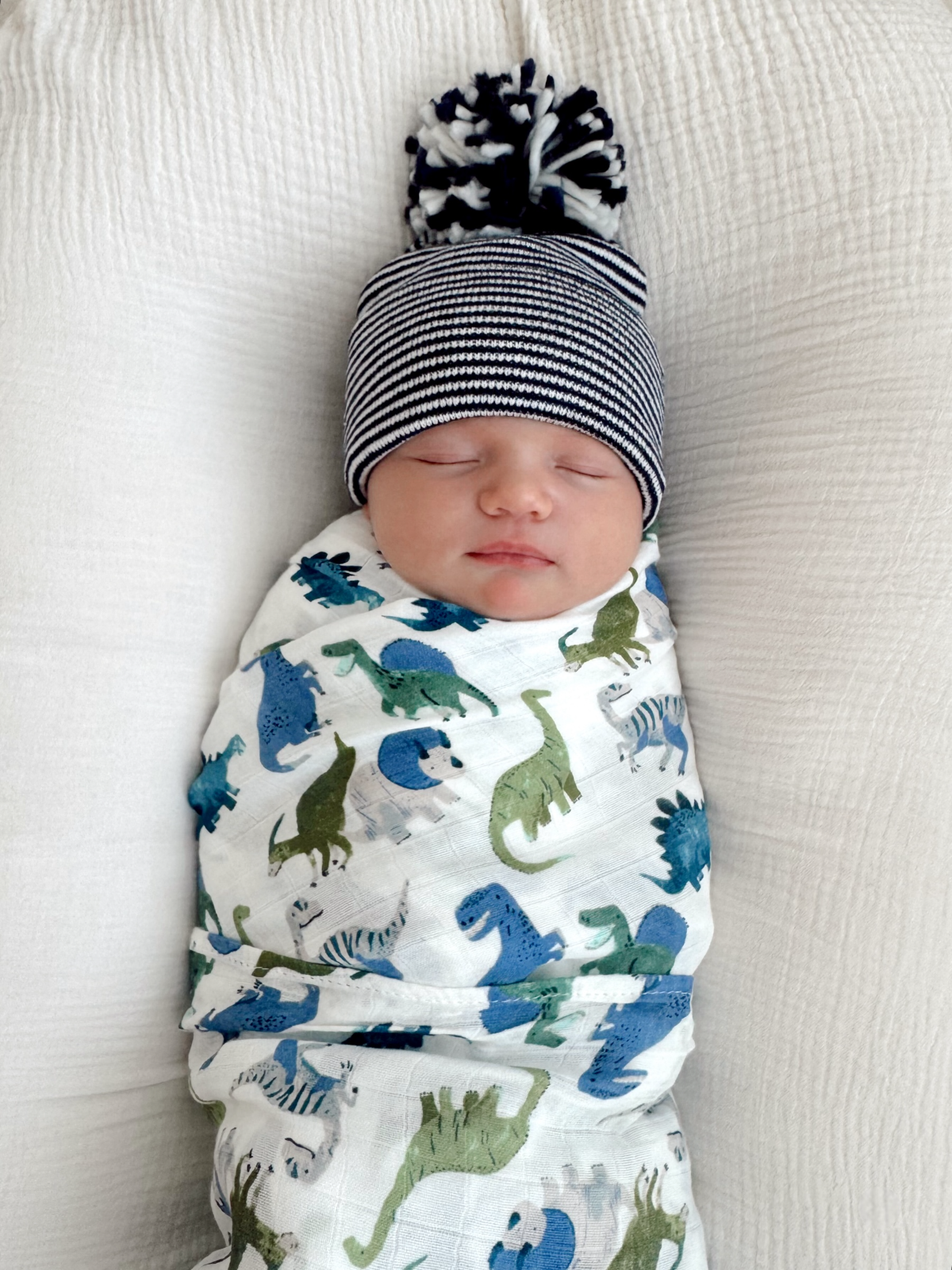 Sleeping baby swaddled in a dinosaur-patterned blanket, wearing a striped beanie with a pom-pom.