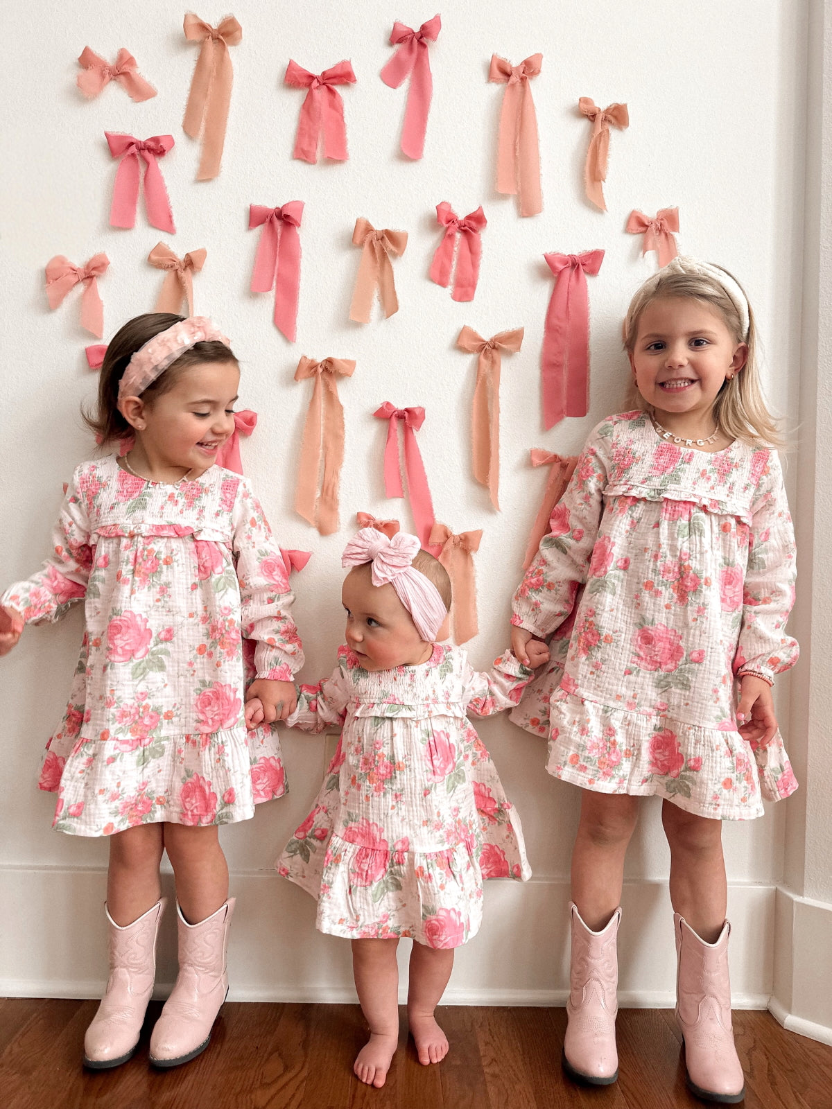 Three young girls in floral dresses holding hands, standing against a wall adorned with pink bows.