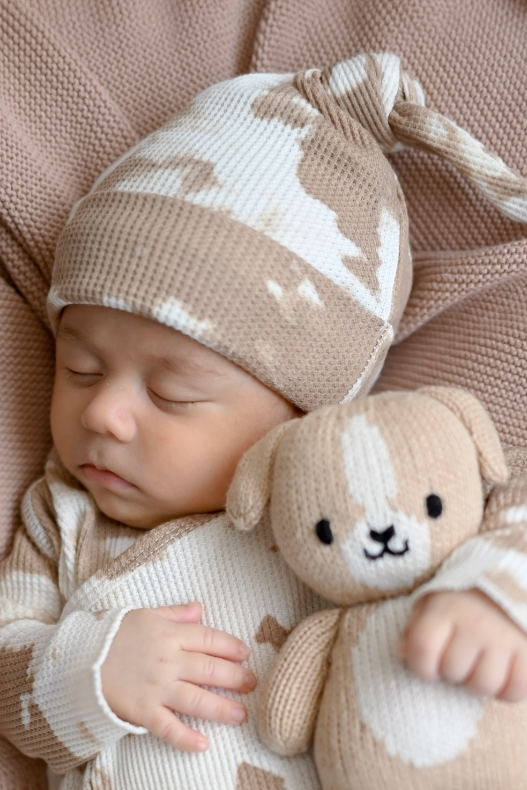 Sleeping baby in a tan outfit, wearing a matching hat, holding a soft toy dog, on a cozy blanket.