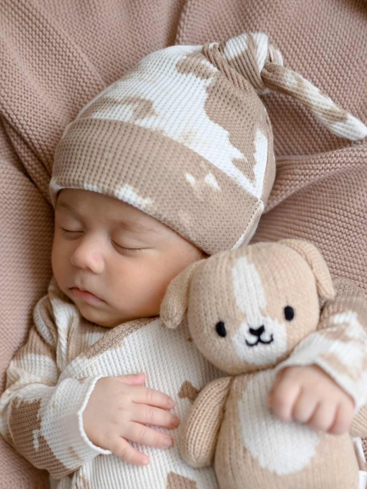 Sleeping baby in a tan outfit, wearing a matching hat, holding a soft toy dog, on a cozy blanket.
